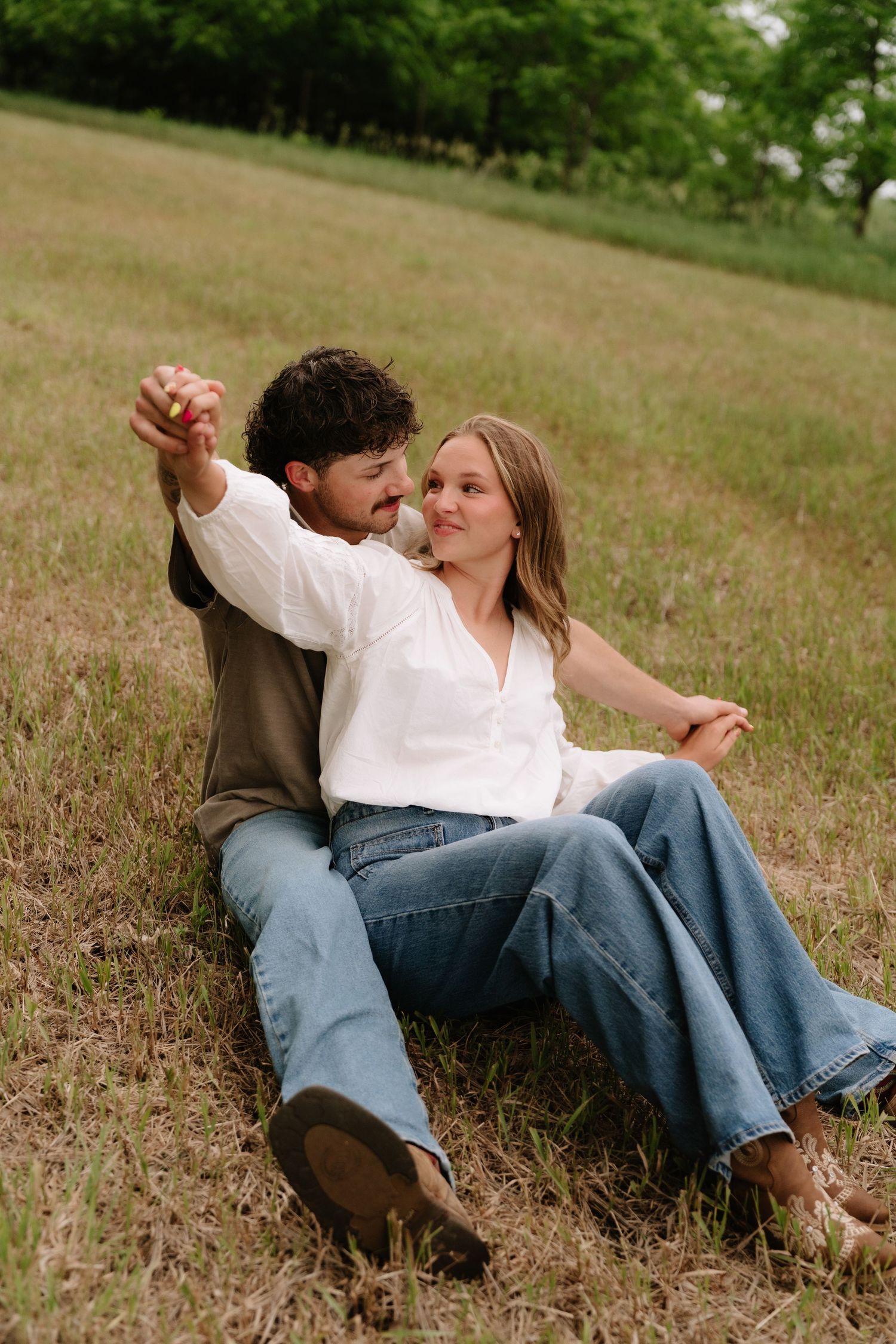 A couple sits together in a grassy field, sharing a tender moment while holding hands outdoors.