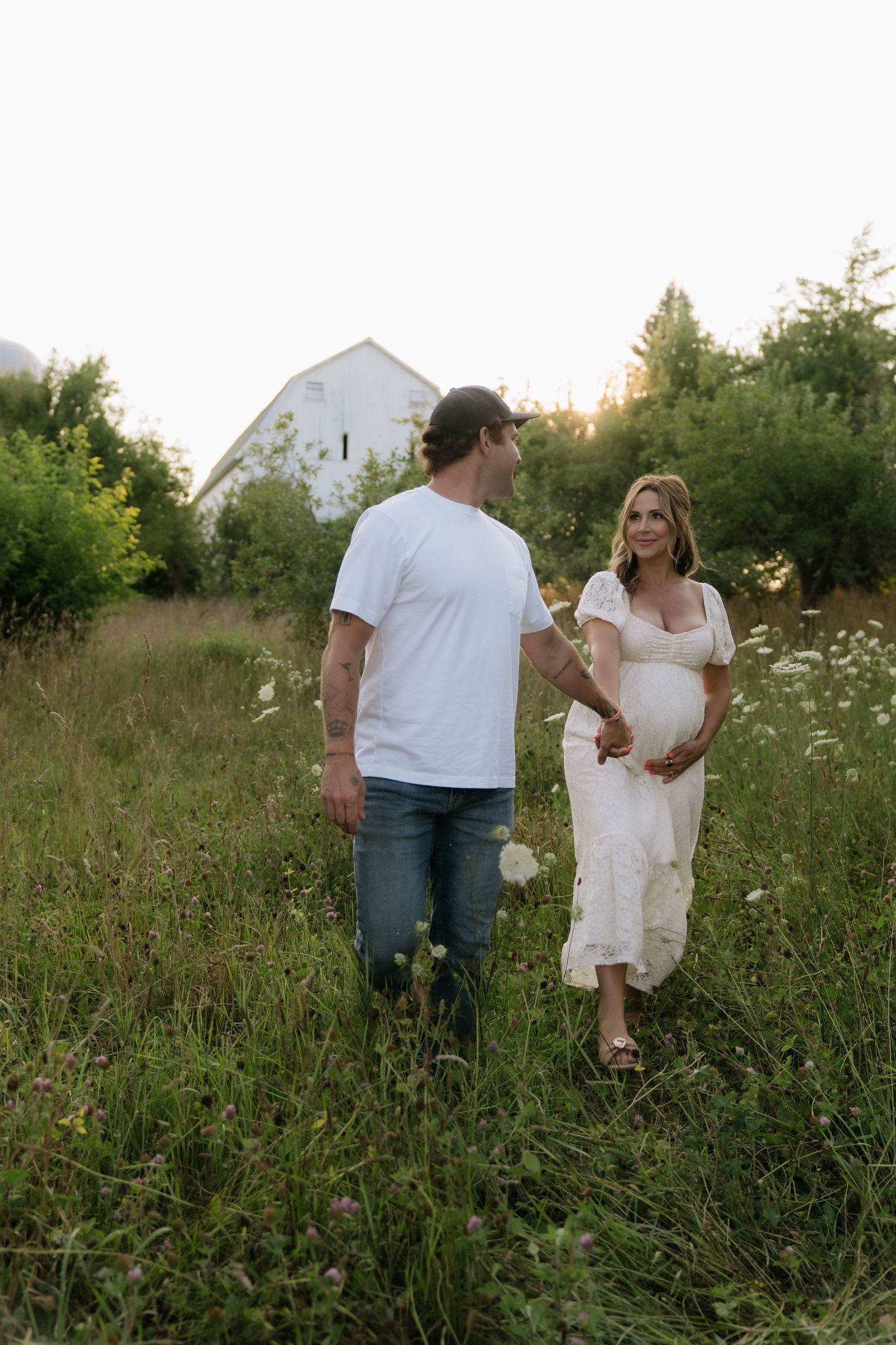 Pregnant couple holds hands walking through a wildflower meadow near a white barn at sunset.
