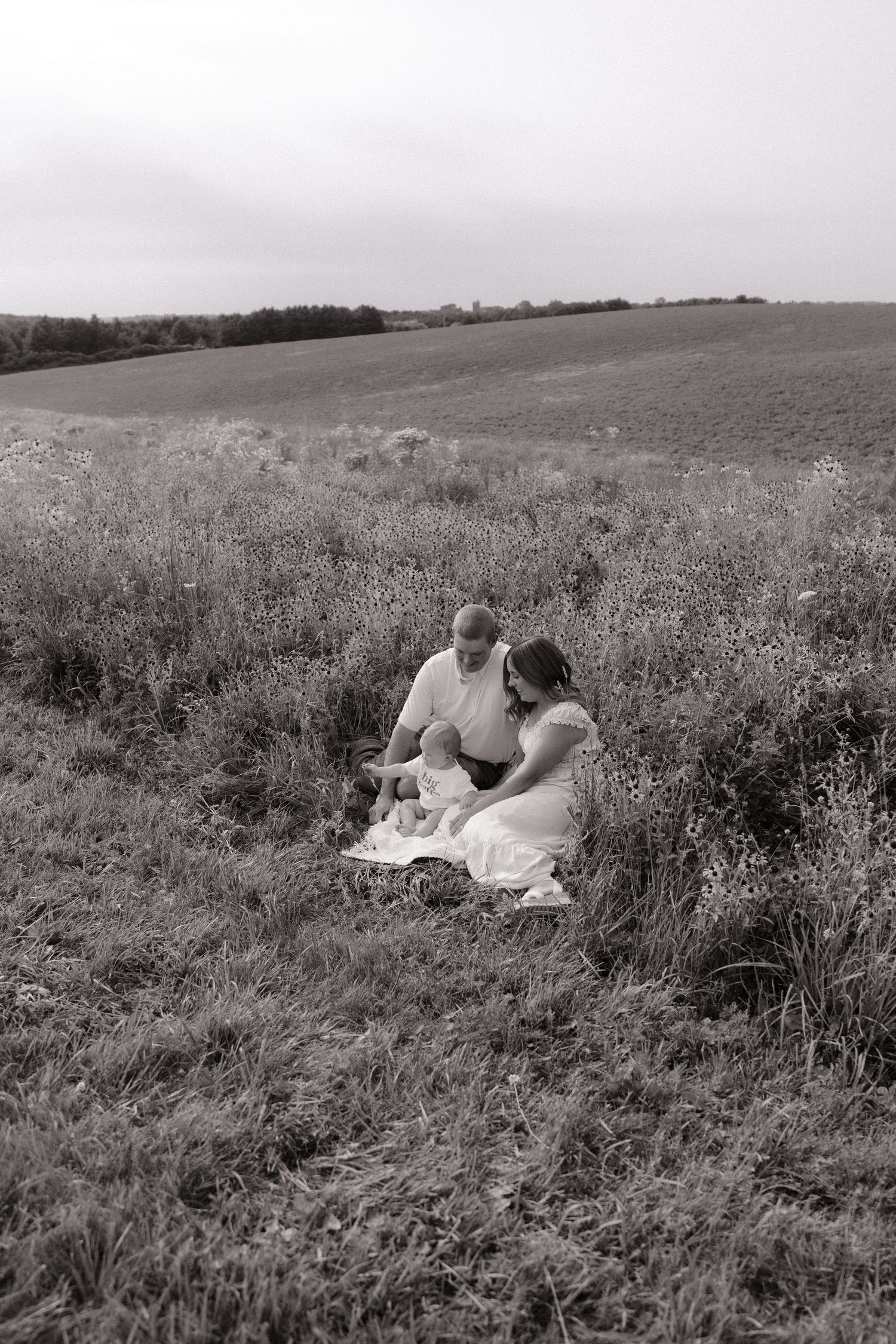Black and white photo of a couple sitting together in a wildflower field, sharing a tender moment outdoors.