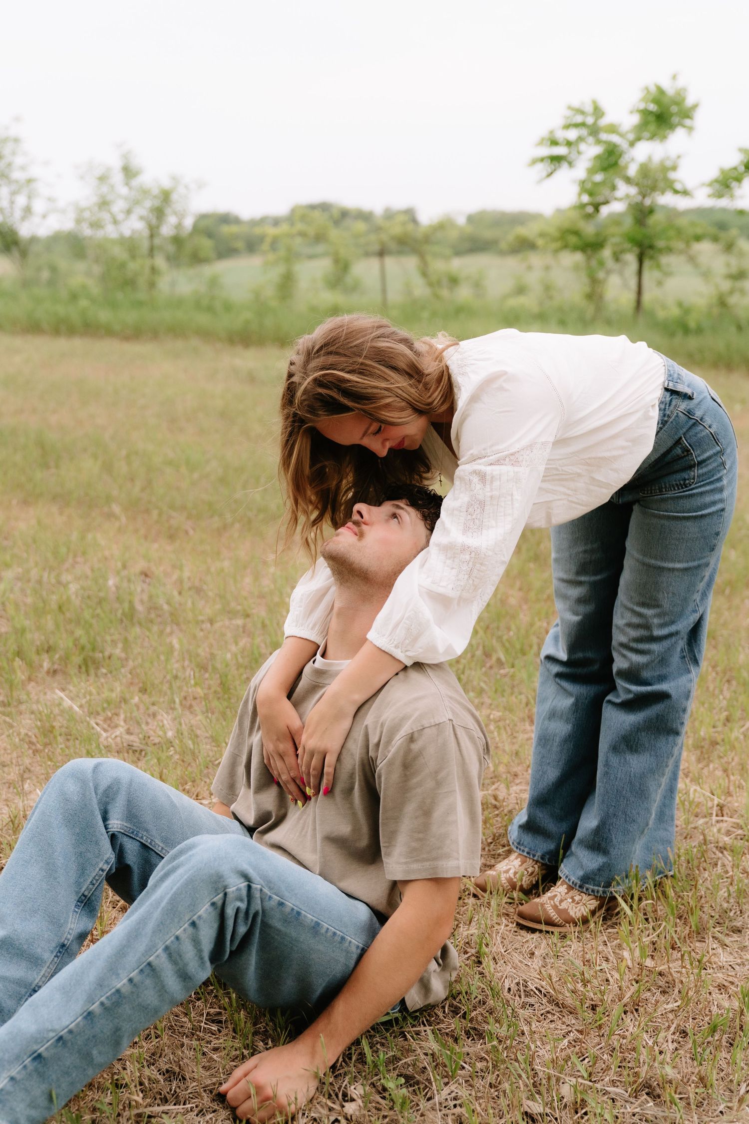 Couple sharing a tender moment outdoors, woman leaning over man sitting on grass in a green field.