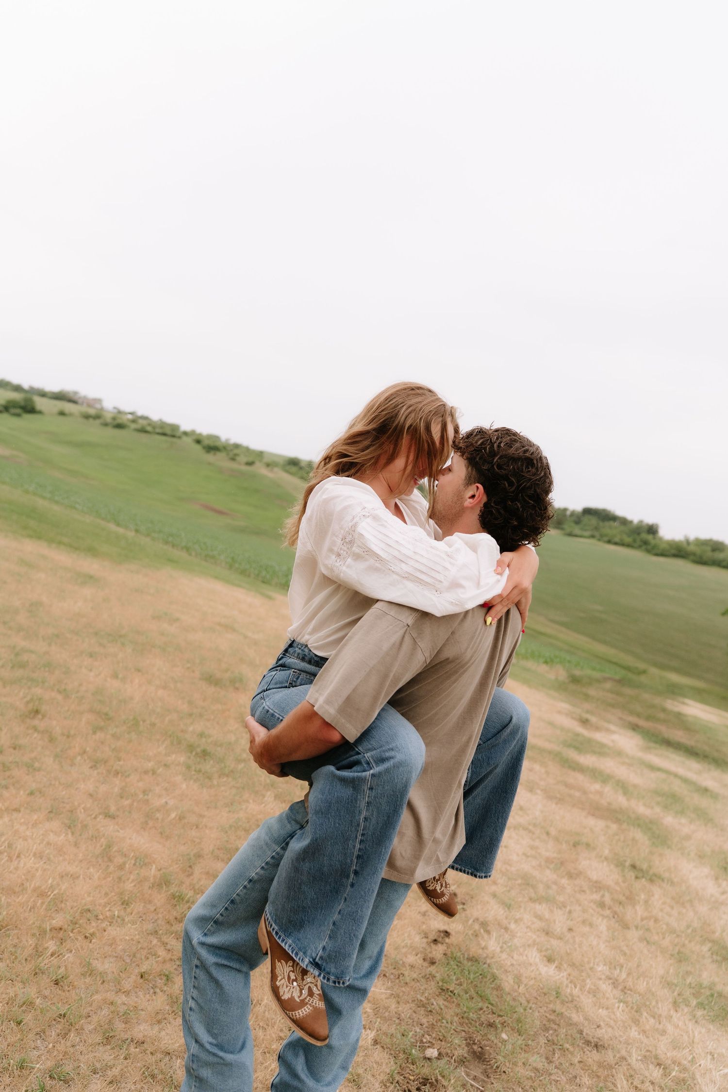 Couple sharing a romantic kiss in an open green field, man lifting woman in casual jeans and white blouse outdoors.