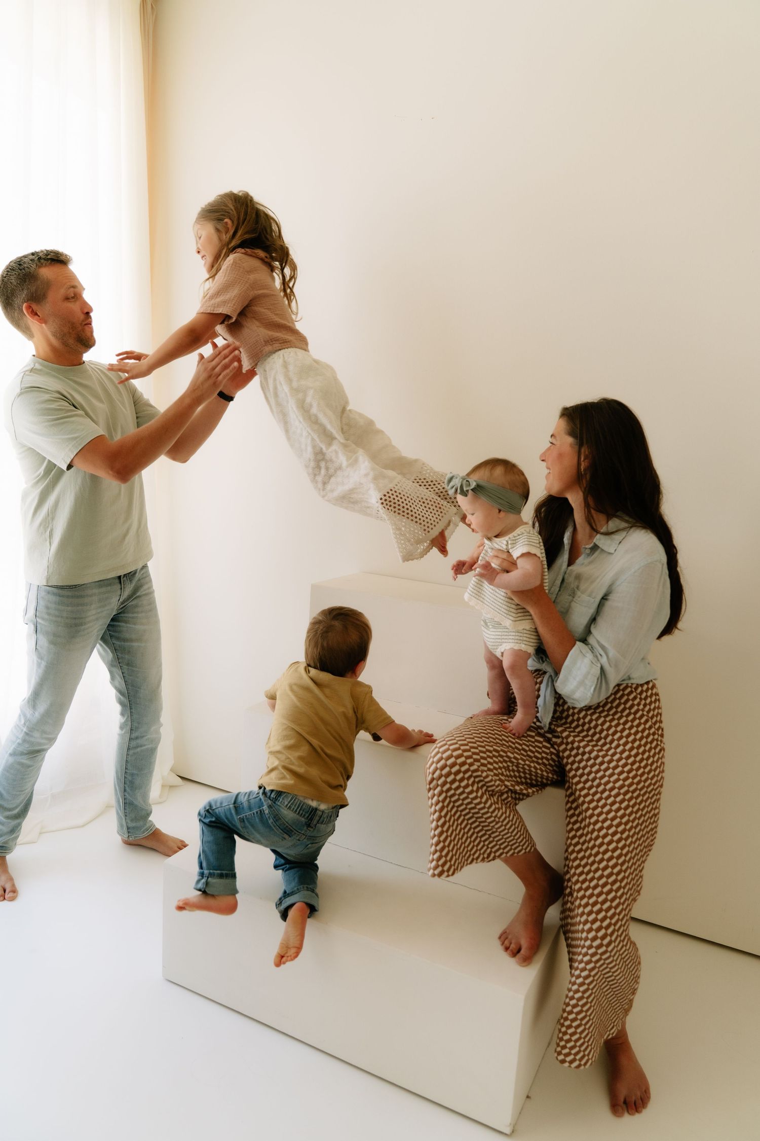A joyful family of five plays together in a bright white studio, with dad lifting a girl while mom holds a baby.
