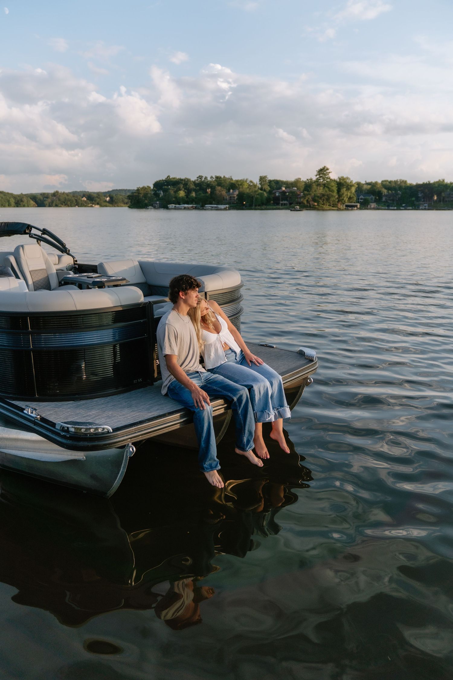 A couple sits closely on the edge of a pontoon boat, sharing a kiss over calm lake waters at golden hour.