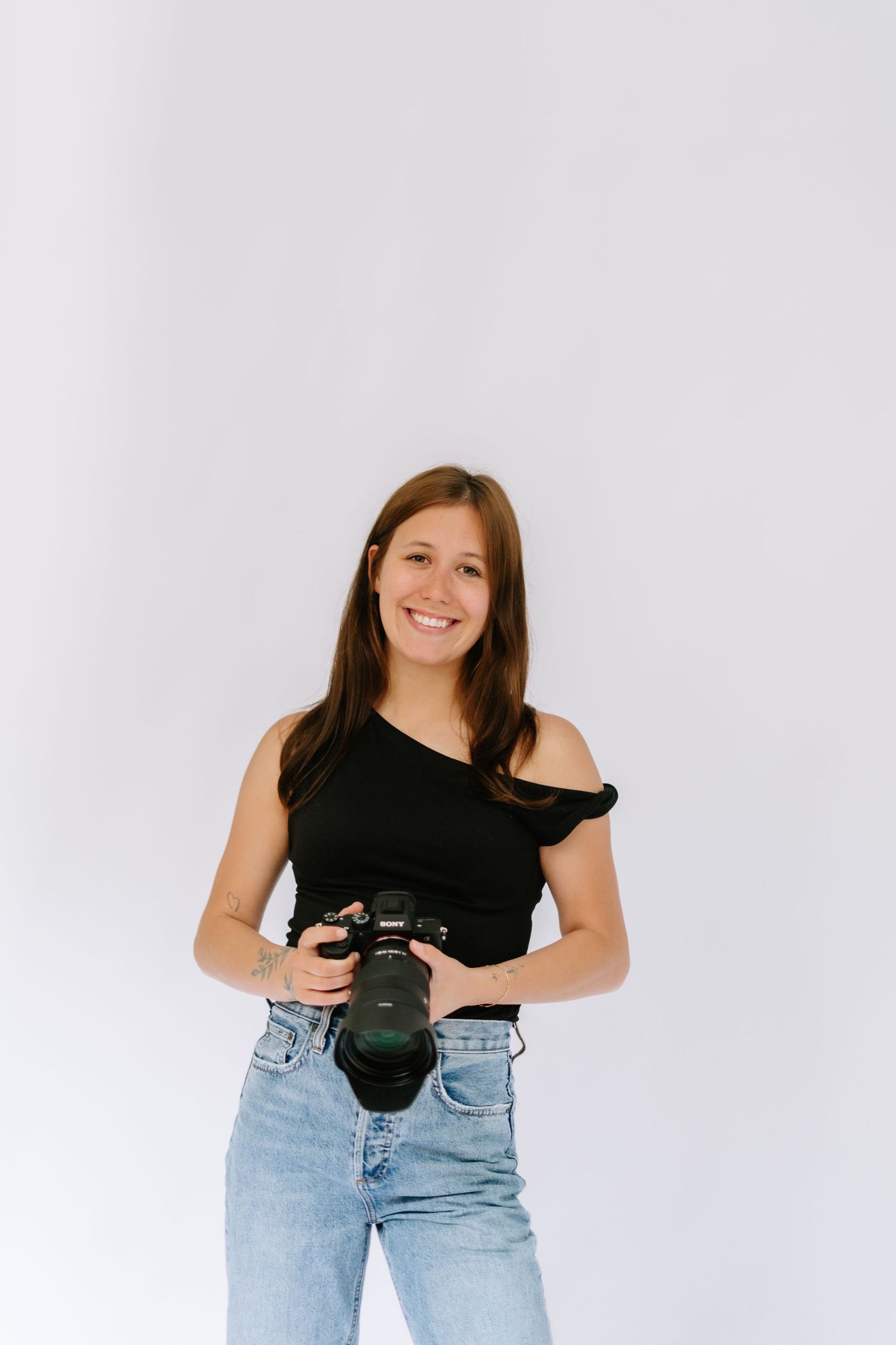 Smiling female photographer in black asymmetric top holding DSLR camera against white background.