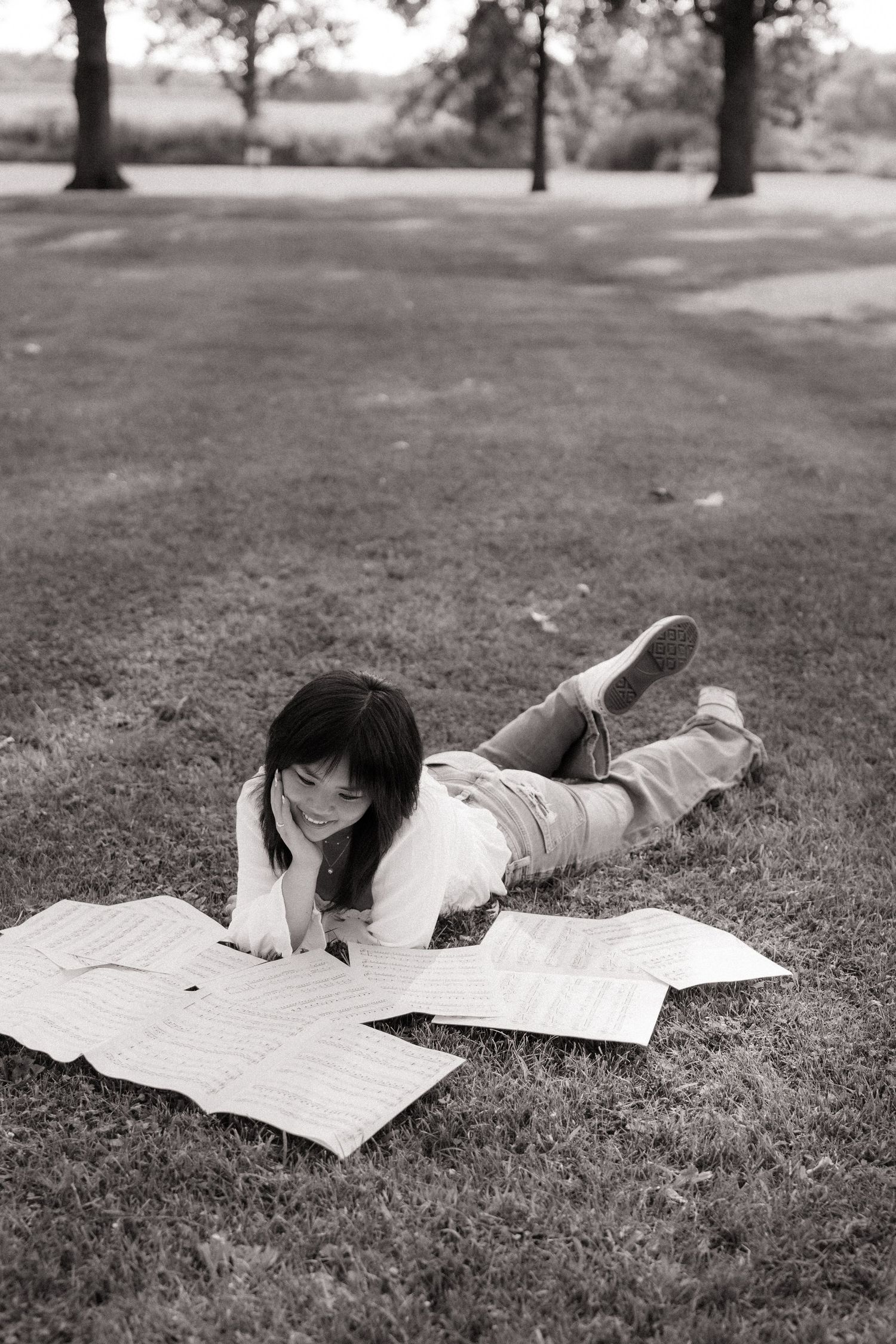 Young woman lying on grass in park studying sheet music papers, black and white photography.