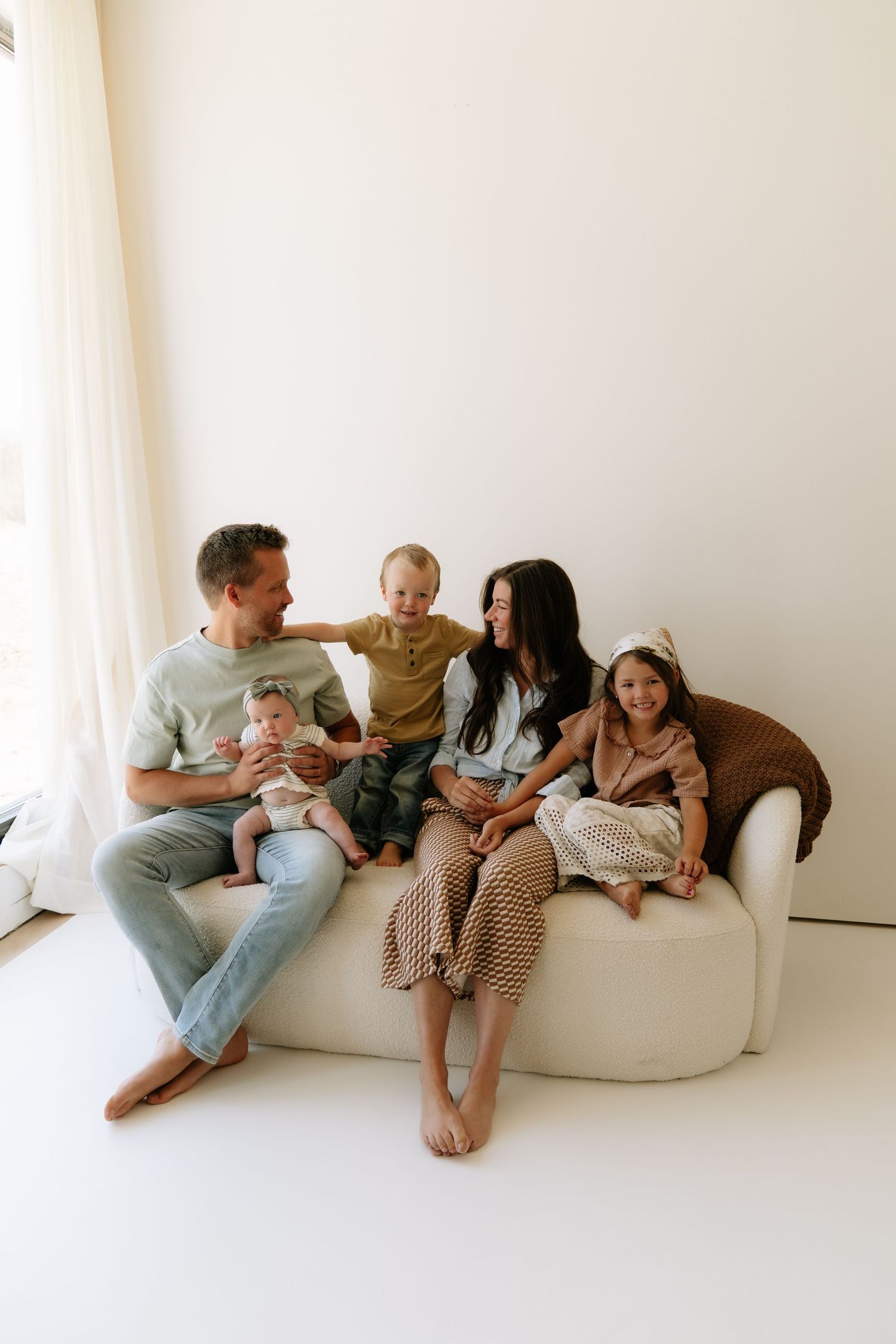 Happy family of five sitting on a cream sofa in a bright white room, parents smiling with three young children.
