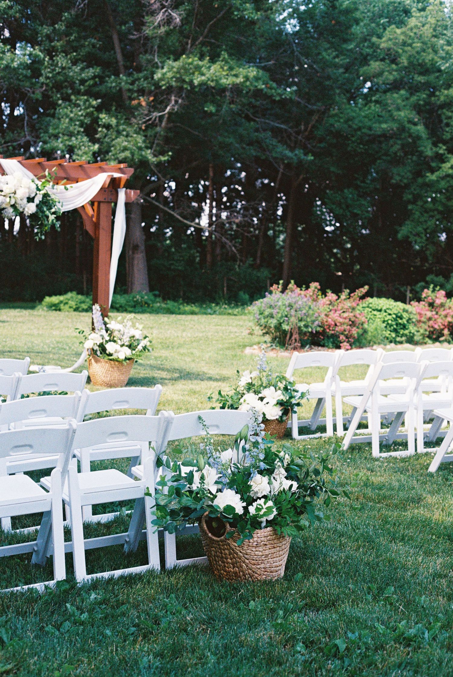 Outdoor wedding ceremony setup with white folding chairs, floral basket arrangements, and a rustic wooden arch on a green lawn.