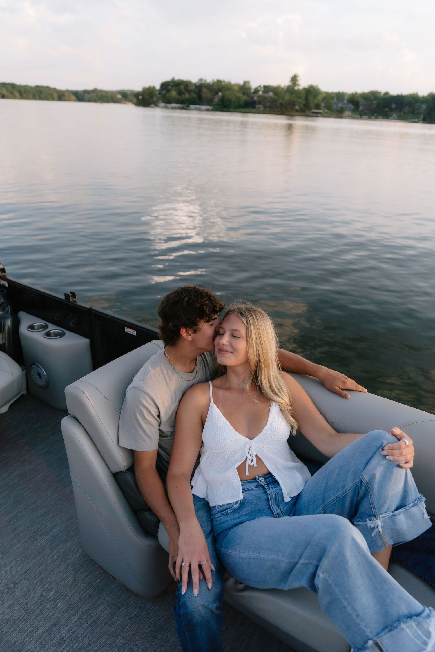 A couple shares a romantic moment on a pontoon boat, with the man kissing the woman's cheek on a calm lake.