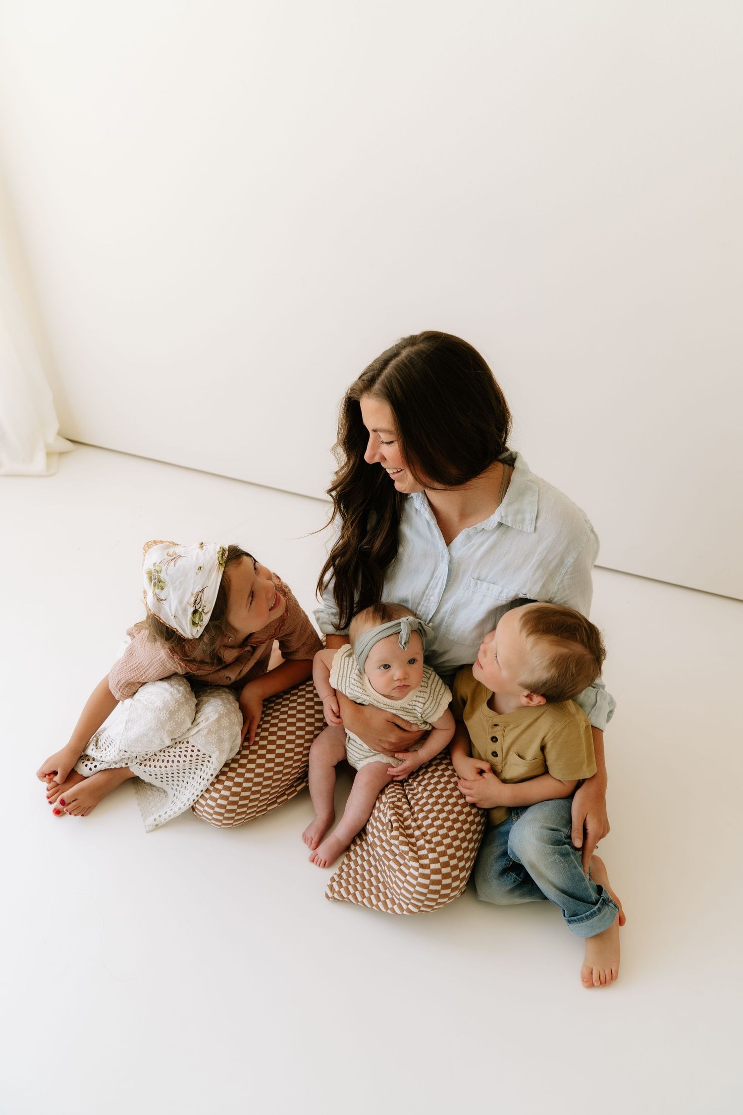A young mother sits on a white studio floor with three young children, smiling warmly in a bright family portrait.