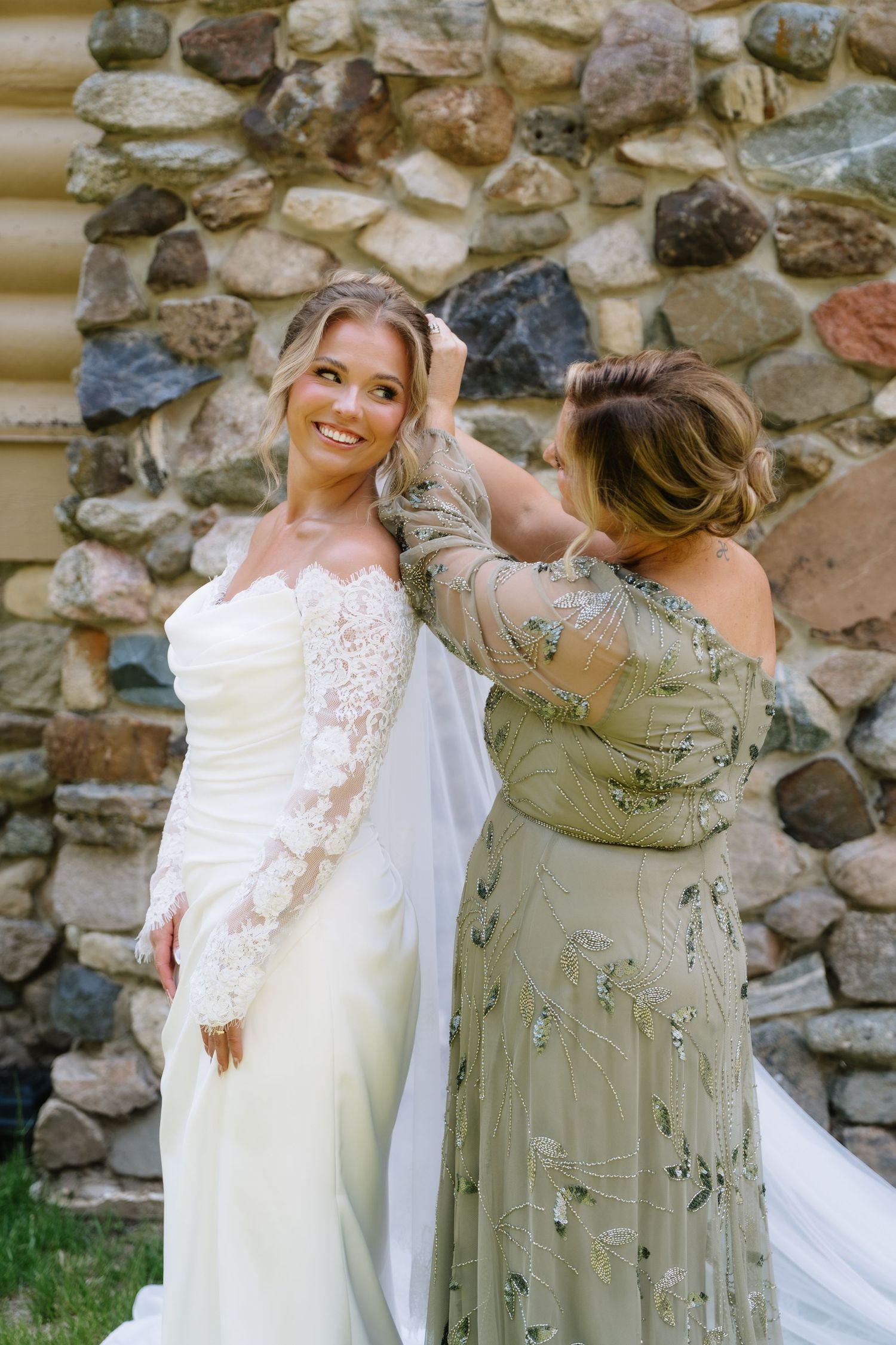 Bride in lace wedding gown smiles as woman in sage green dress adjusts her veil near stone wall.