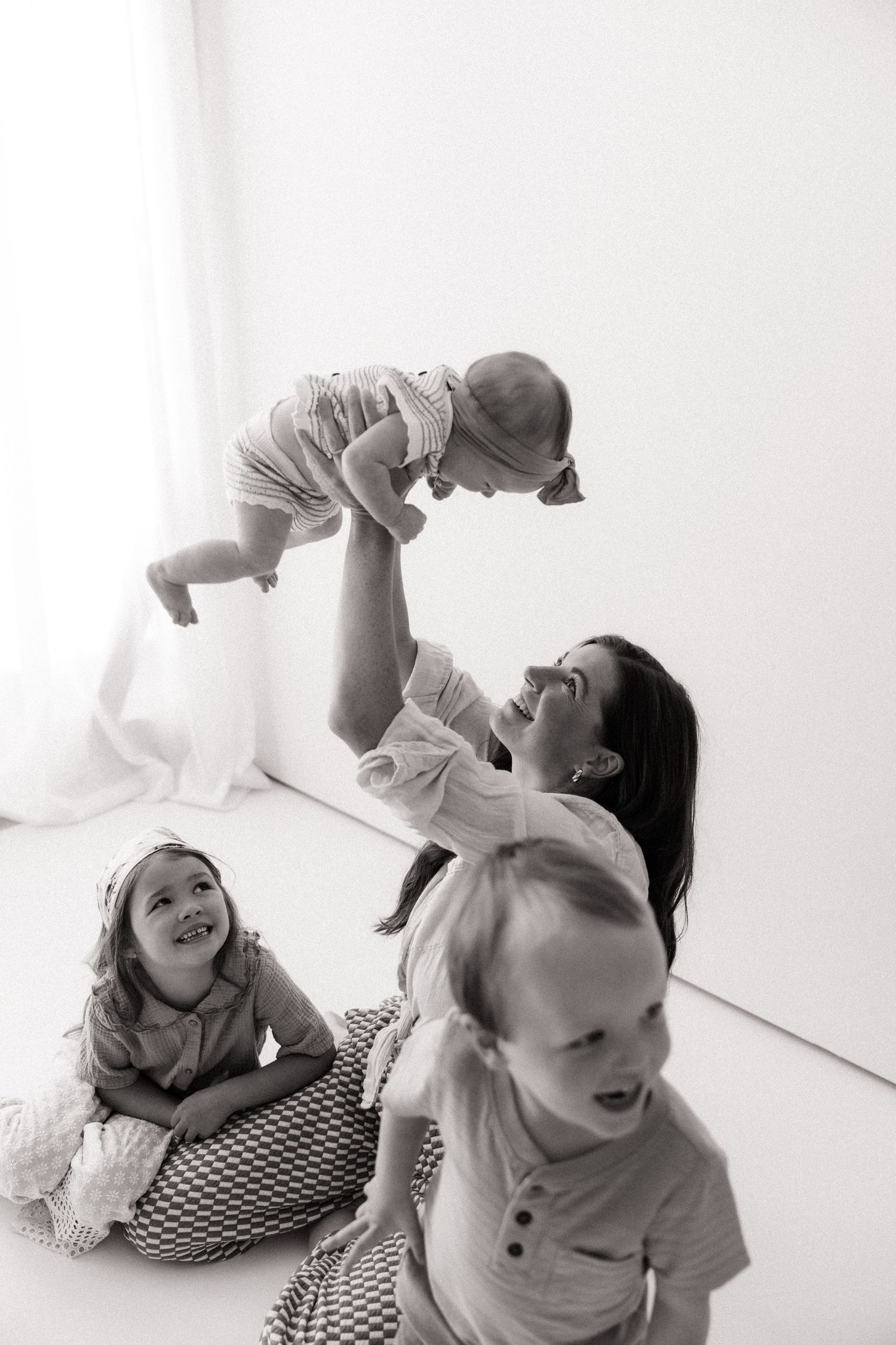 Black and white photo of a mother lifting her baby in the air while two young children sit nearby on the floor.