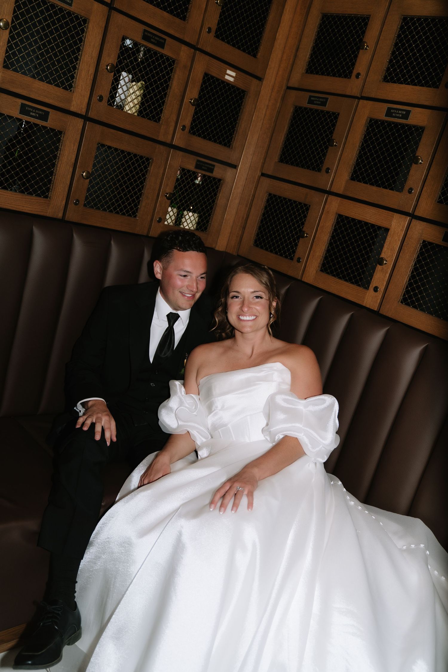 A smiling bride in a white off-shoulder gown and groom in black tuxedo sit together on a leather booth seat.