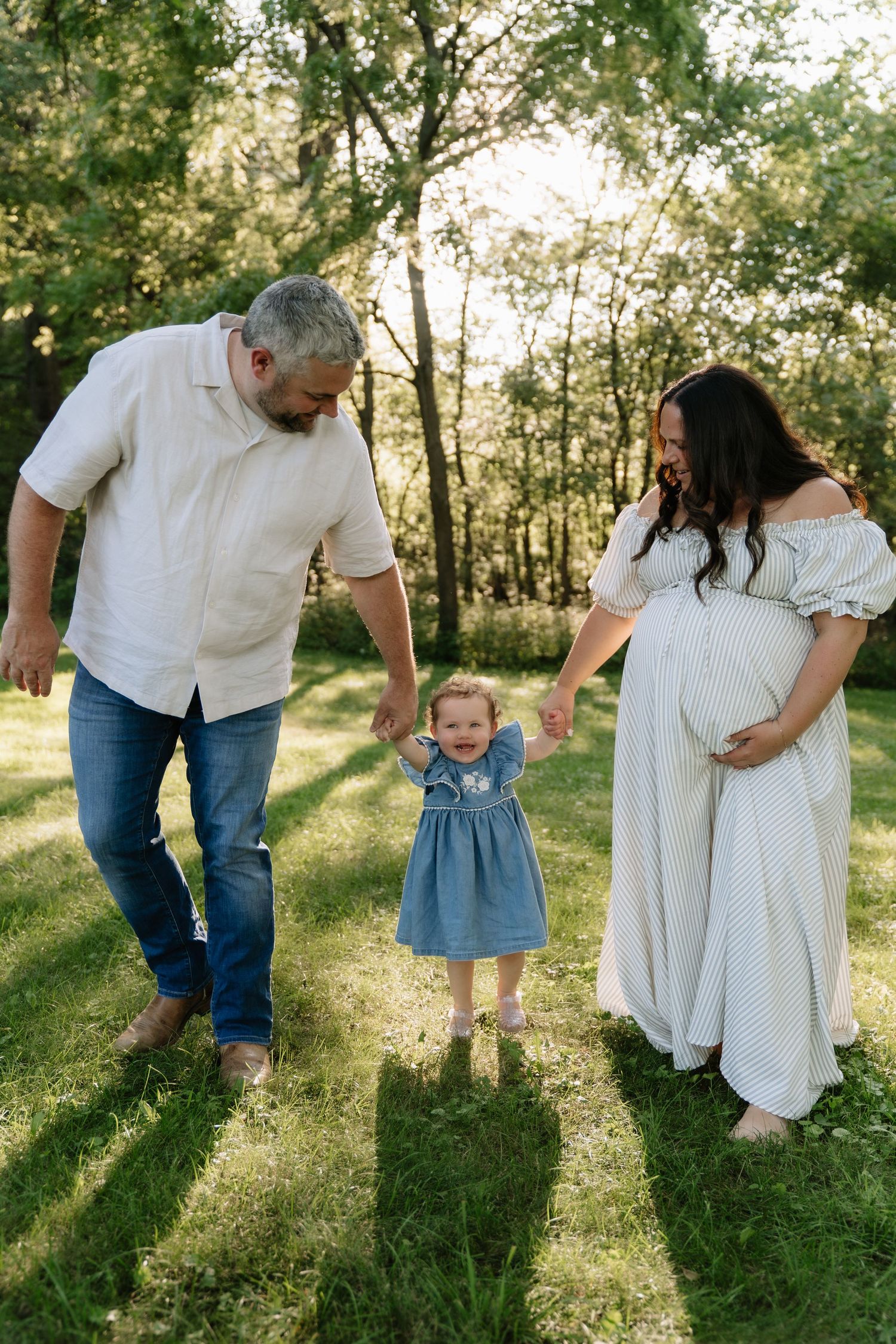 Pregnant woman in white dress holds hands with toddler girl and husband in sunny green park during maternity photo session.