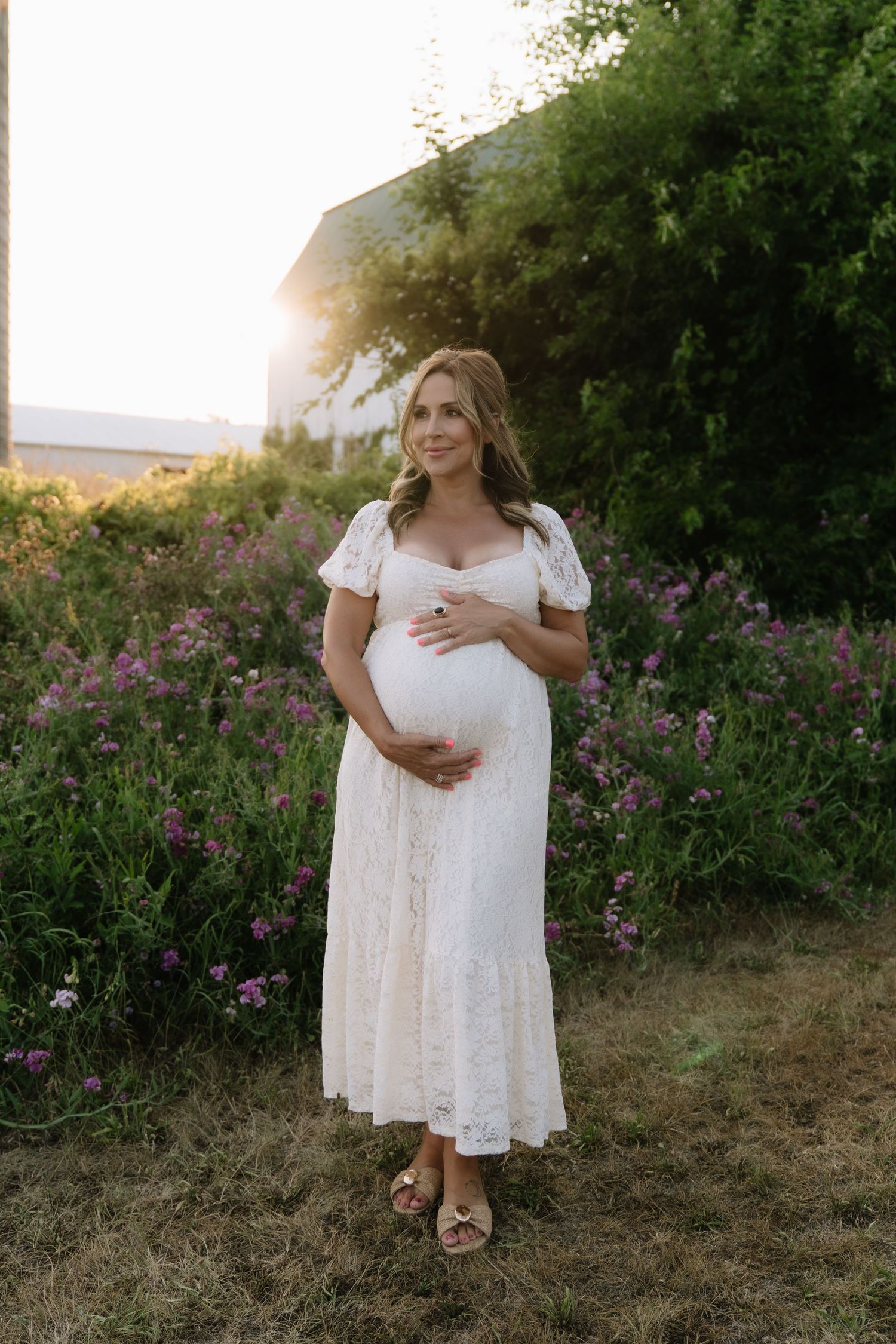 Pregnant woman in white lace dress holding baby bump in garden at golden hour sunset.