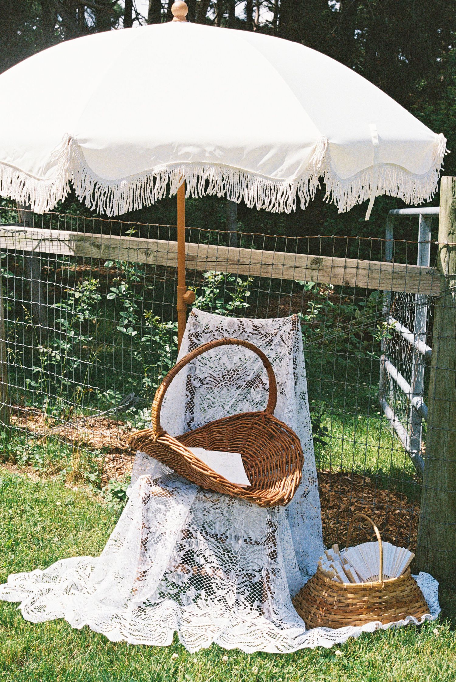 Vintage wicker baskets on lace tablecloth under white fringed beach umbrella at outdoor garden wedding.