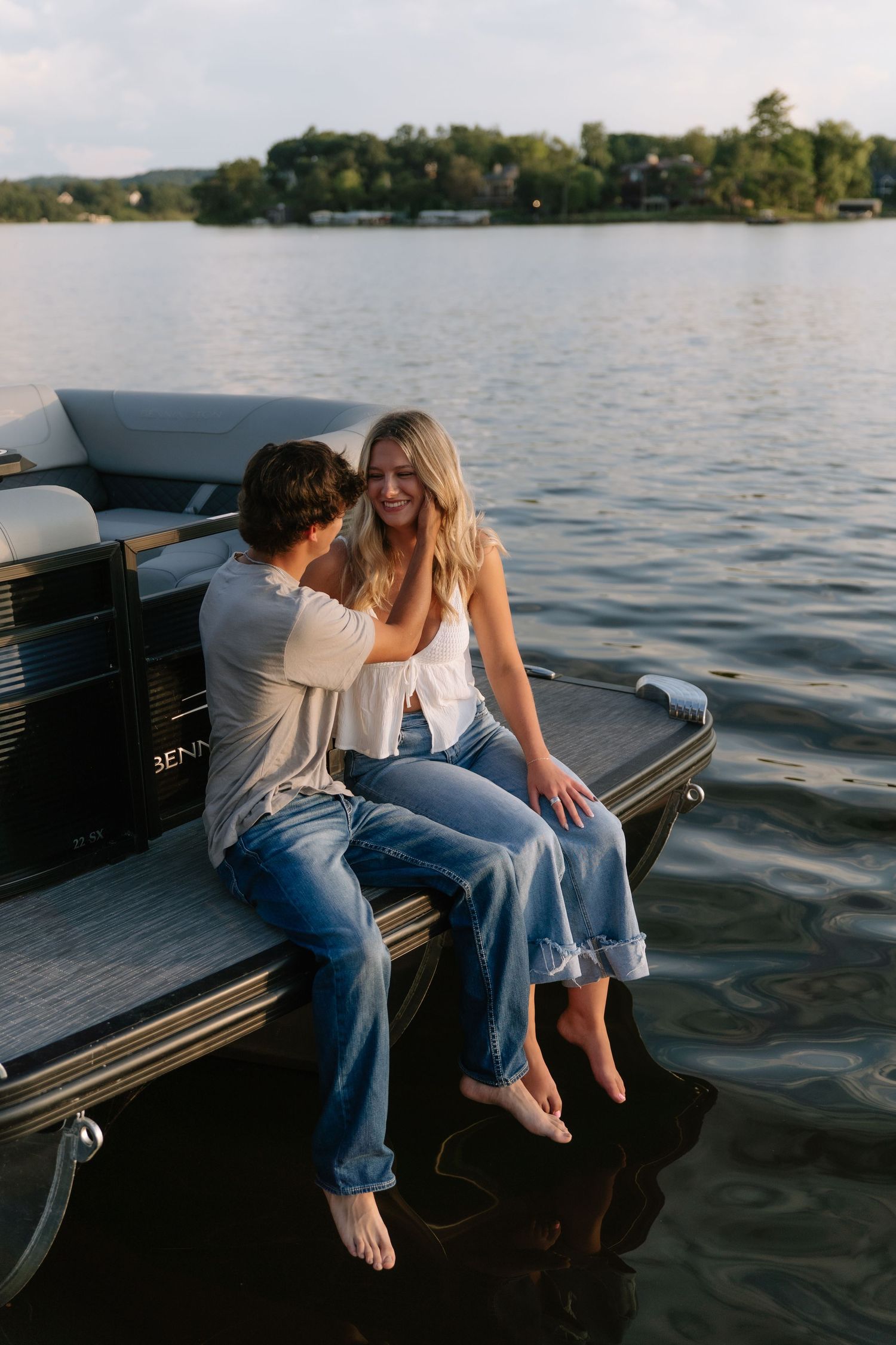 Couple sitting on the edge of a pontoon boat, smiling and talking together on a calm lake at sunset.