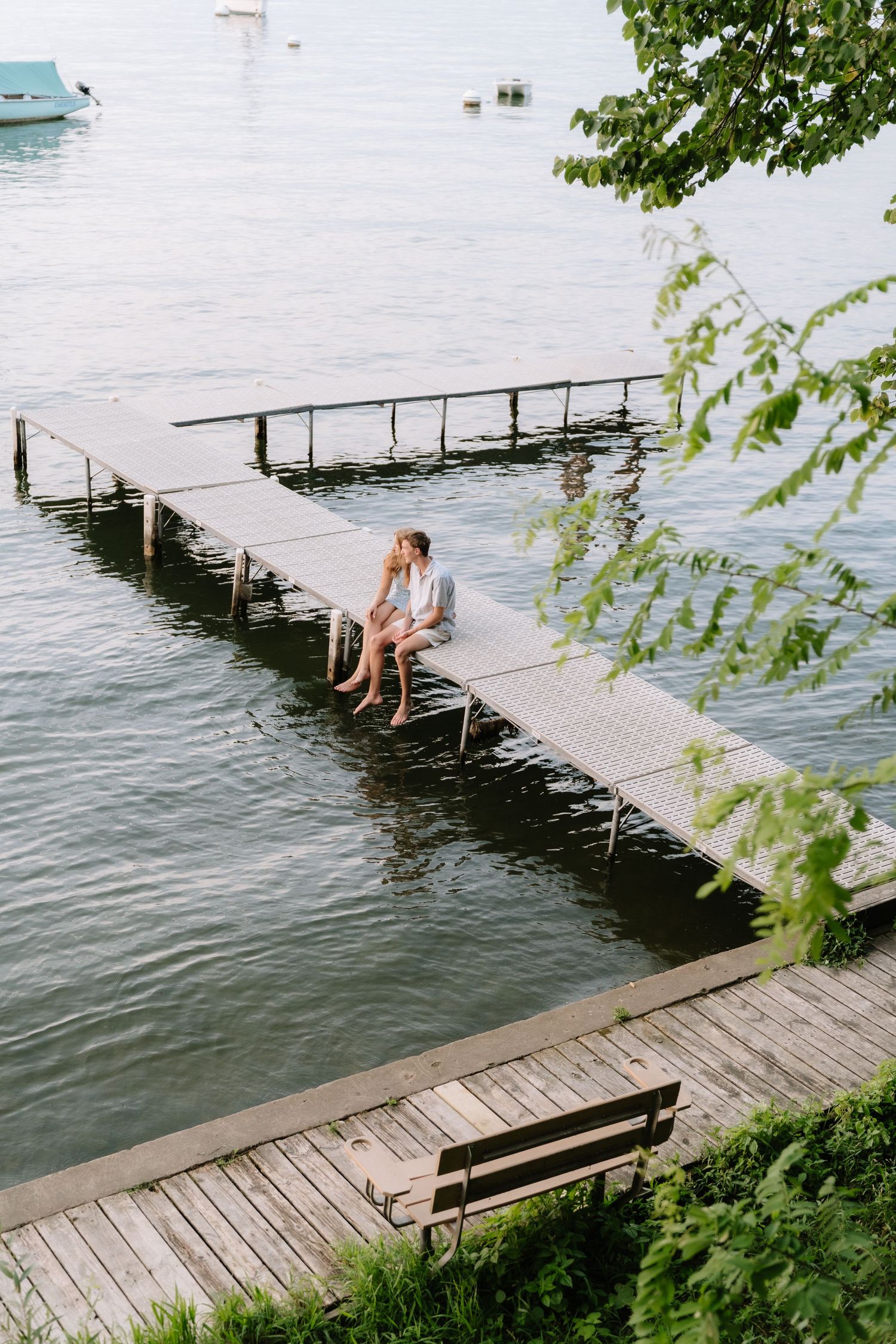 Couple sitting close together on a wooden dock over calm lake water, with a bench and green trees nearby.