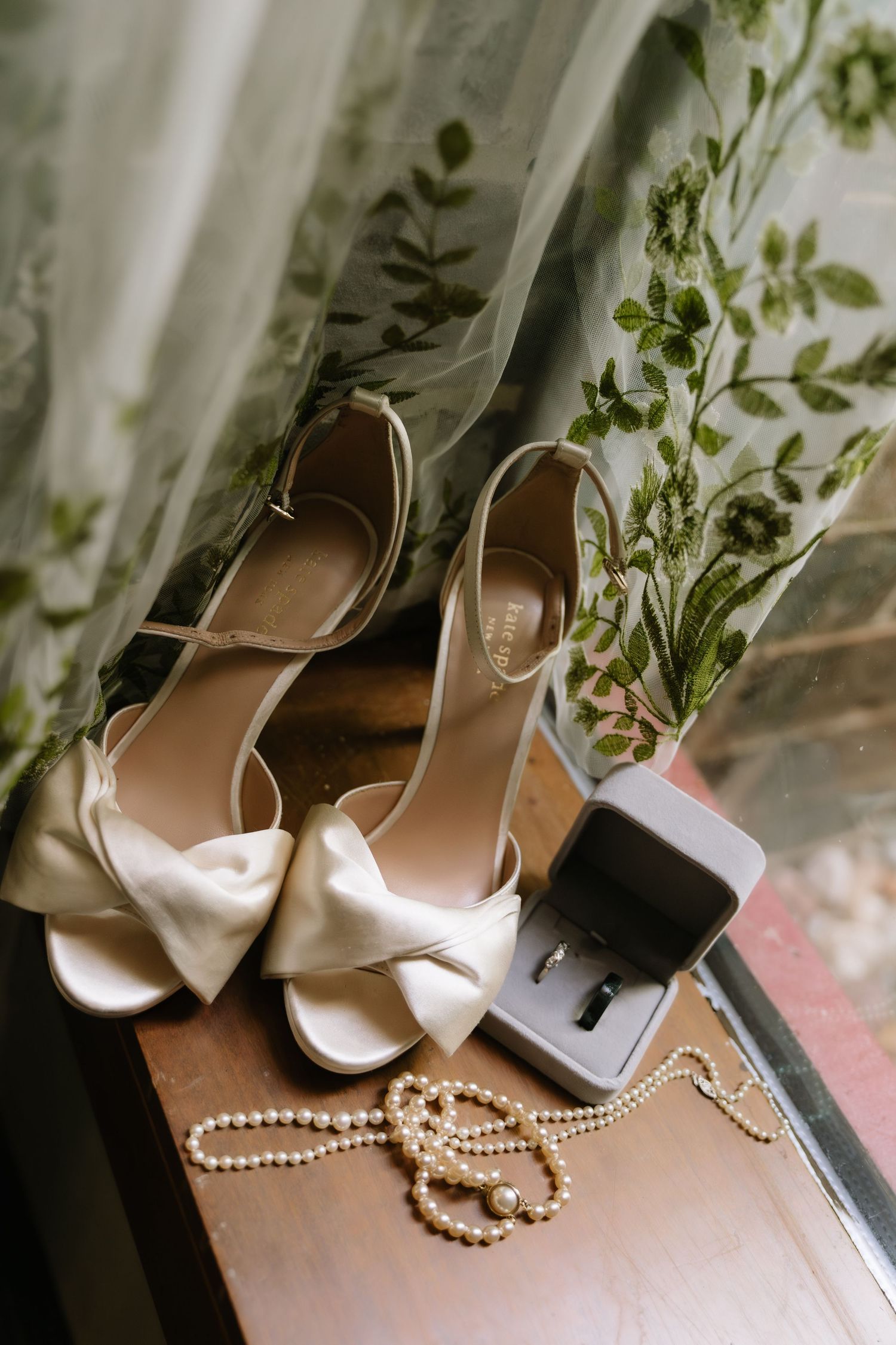 Elegant white bridal heels with bow detail displayed with pearl necklace and ring box.