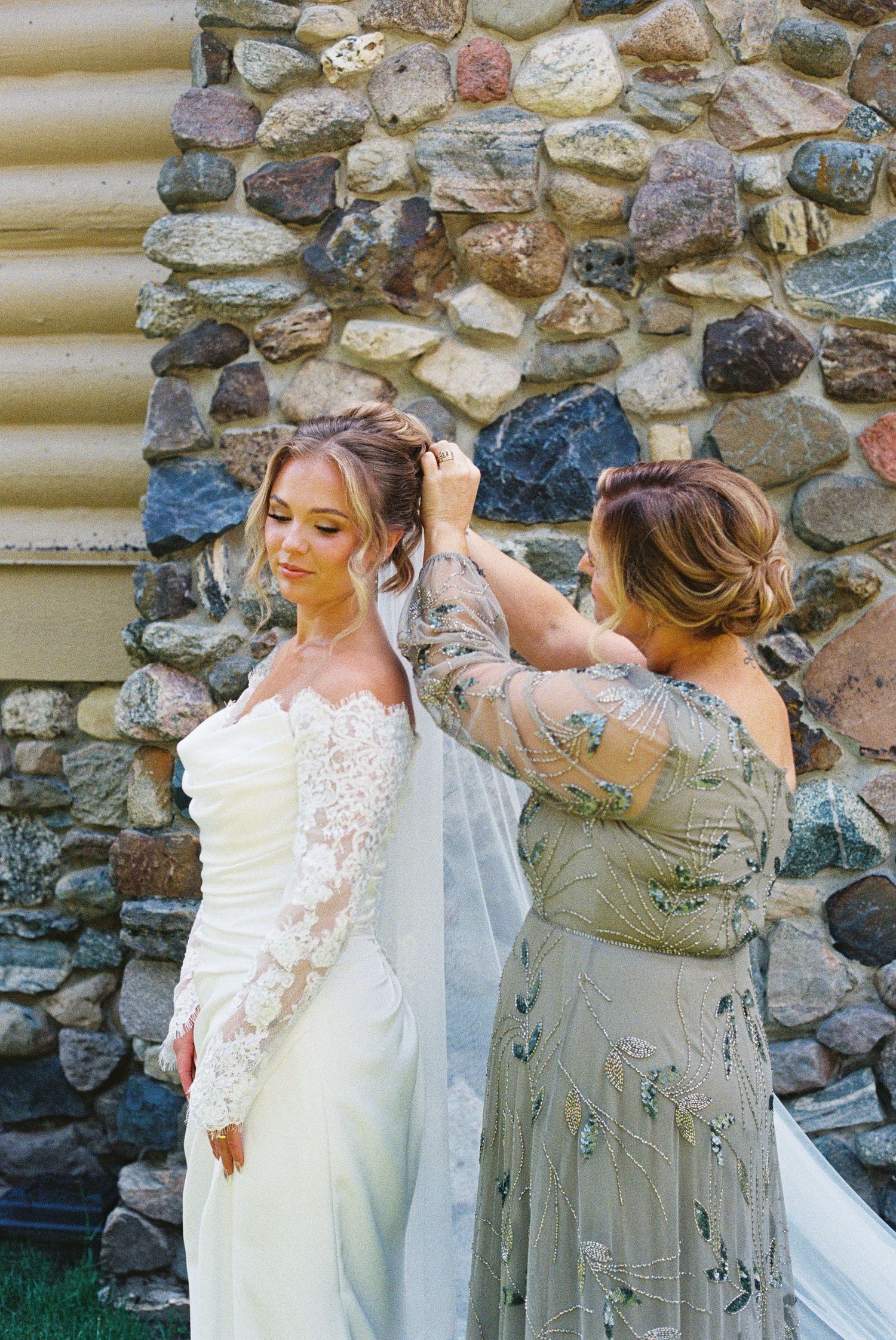 Bride in lace wedding gown having veil adjusted by bridesmaid in floral dress against stone wall backdrop.