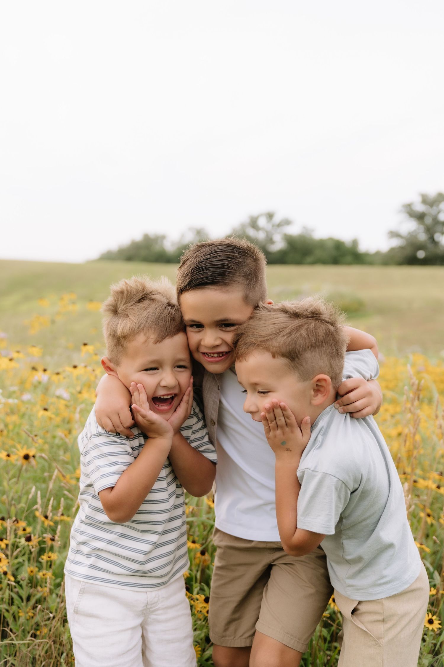 Three young boys hugging and laughing together in a field of yellow wildflowers on a cloudy day.