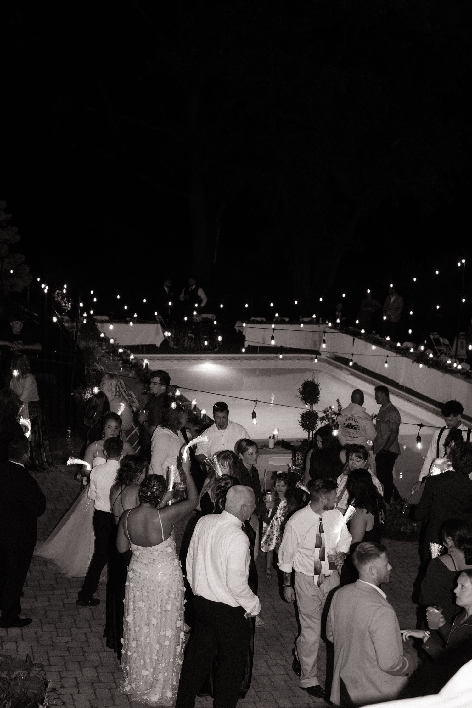 Black and white photo of wedding guests dancing outdoors at night under string lights at an elegant evening reception.