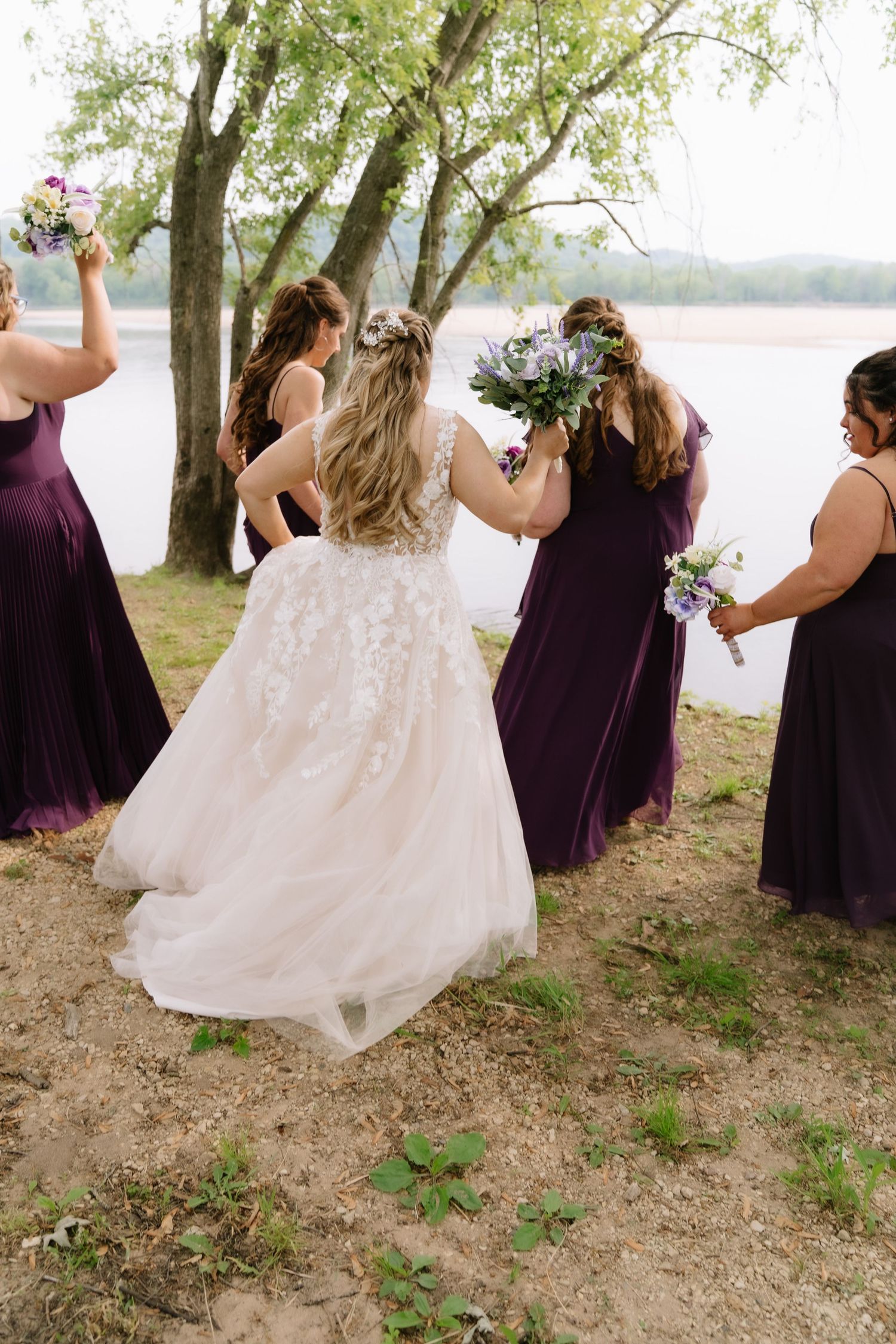 Bride in lace gown with bridesmaids in purple dresses holding bouquets near a lakeside tree.