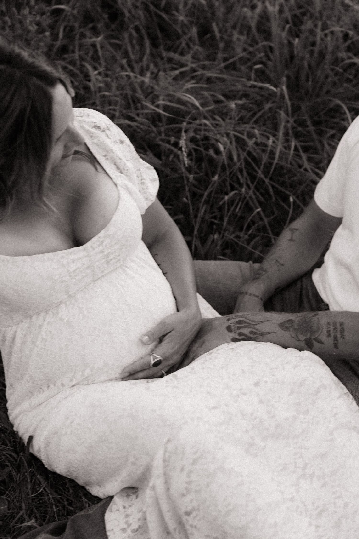 Black and white maternity photo of tattooed couple sitting in tall grass, hands on baby bump.