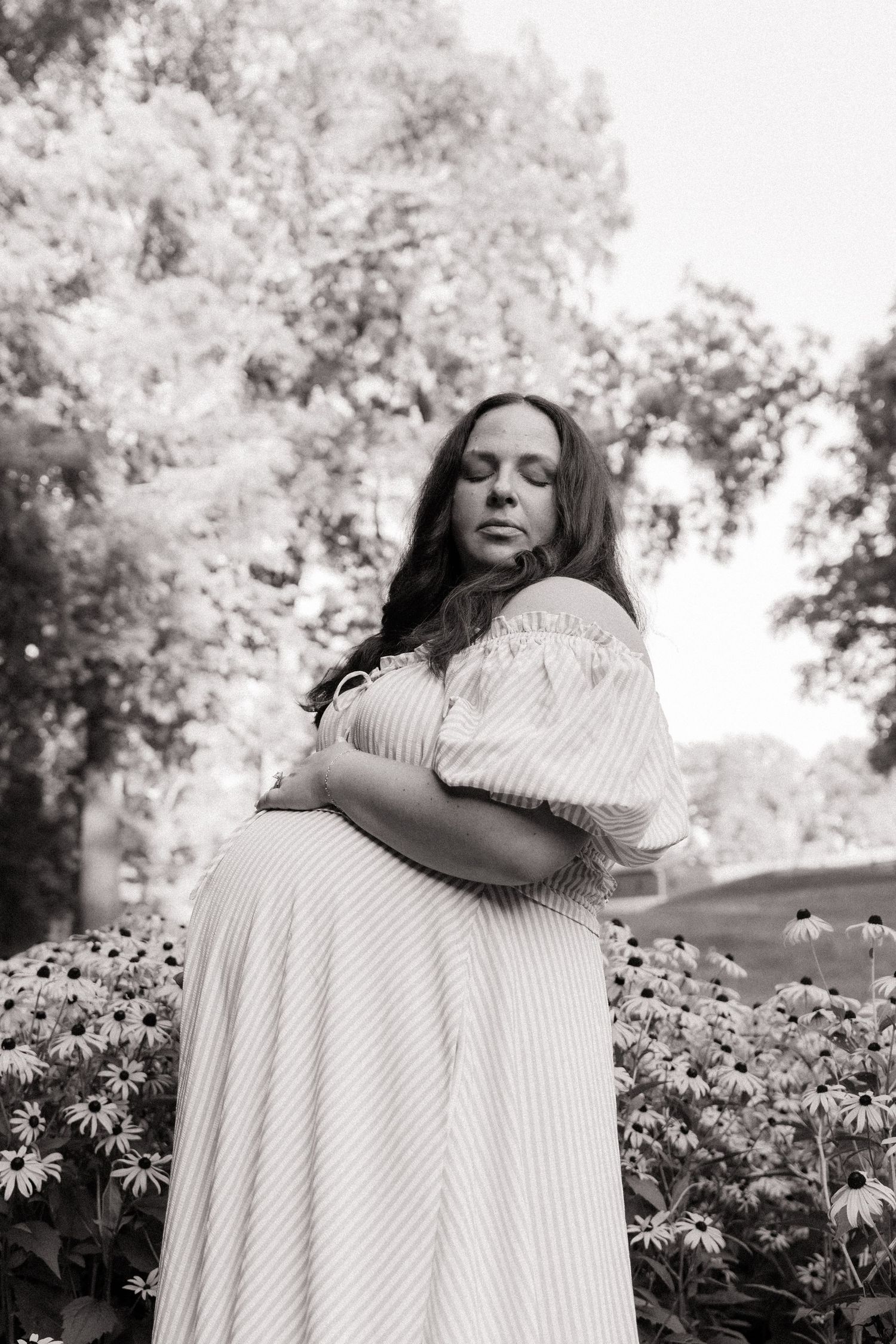 Black and white photo of a pregnant woman in a striped dress cradling her belly among flowers in a lush garden.
