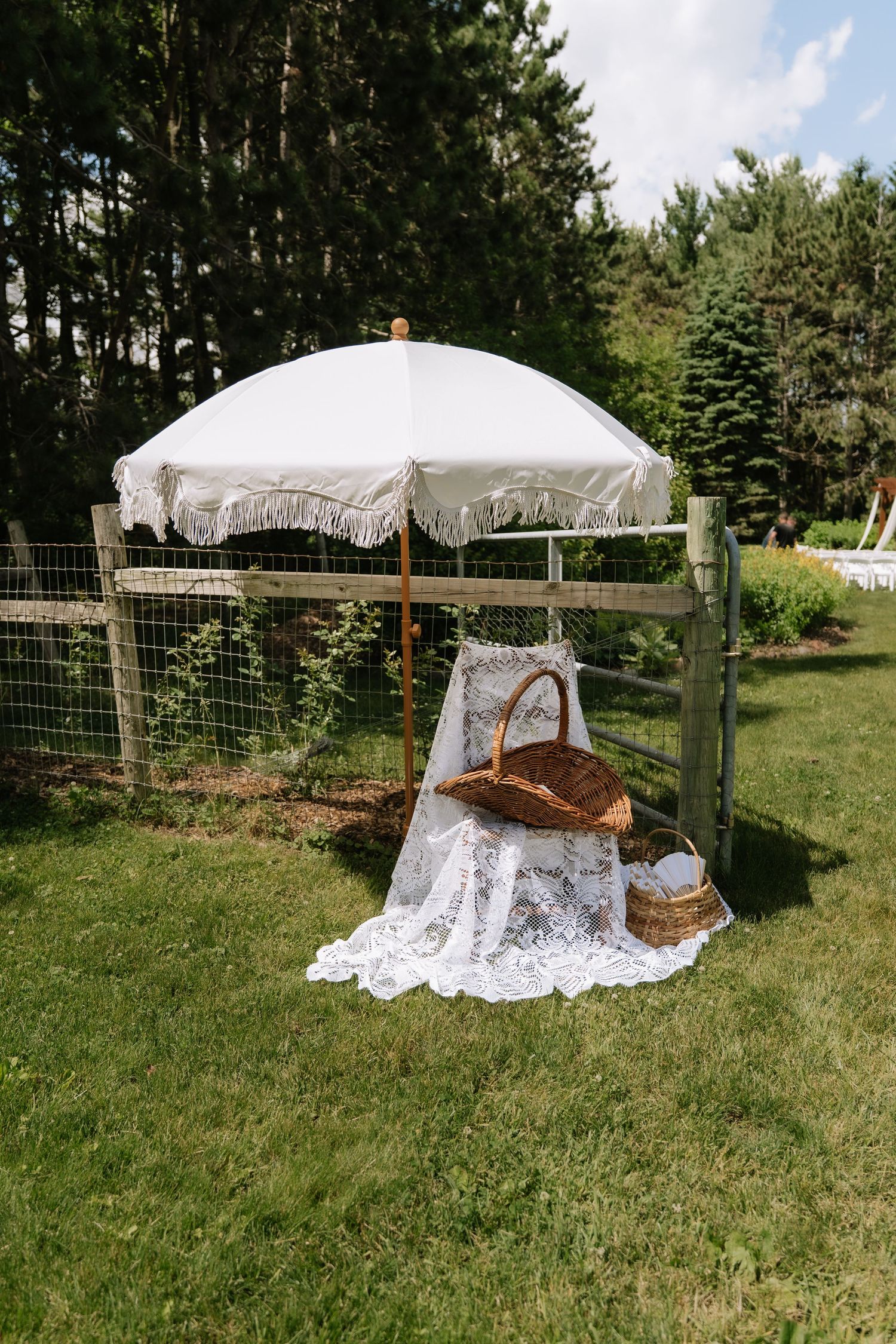 White fringed patio umbrella shading wicker picnic baskets on lace cloth in a lush green garden setting.