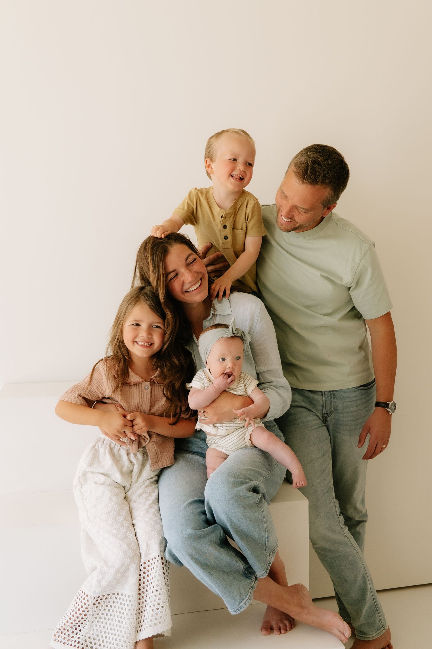 Happy family of five with mom, dad, toddler, young girl, and newborn baby posing together in a bright white studio.