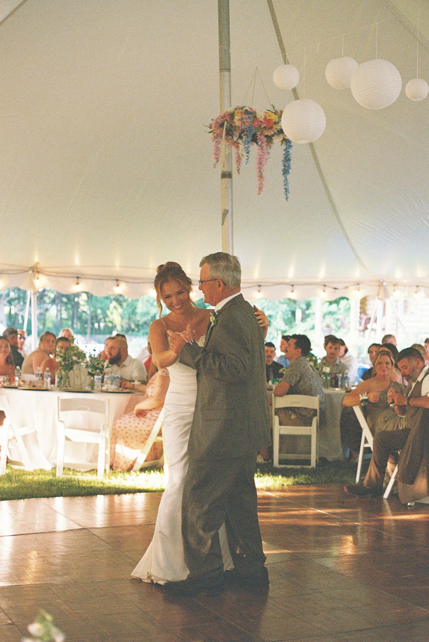 Bride and father share an emotional first dance under a white tent with floral chandelier at an outdoor wedding reception.