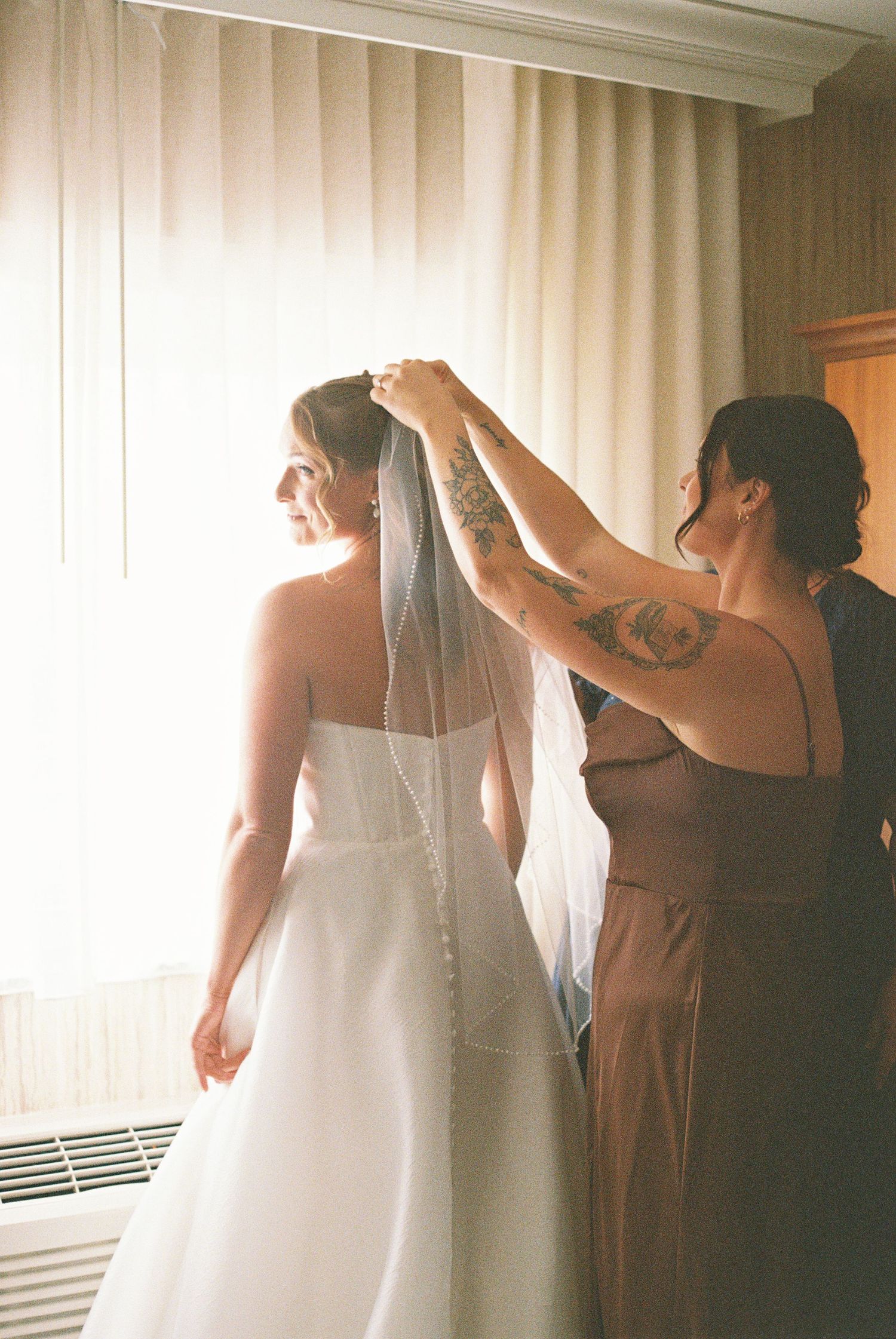 A bridesmaid with tattoos adjusts the veil of a bride in a white strapless gown near a sunlit window.