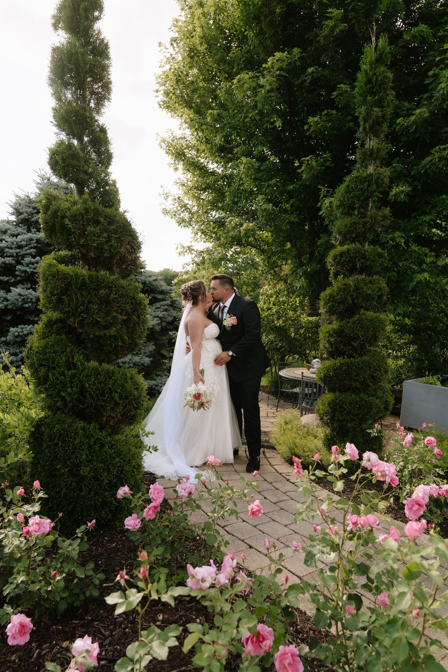 A bride and groom share a romantic moment on a brick path surrounded by lush greenery and blooming pink roses.