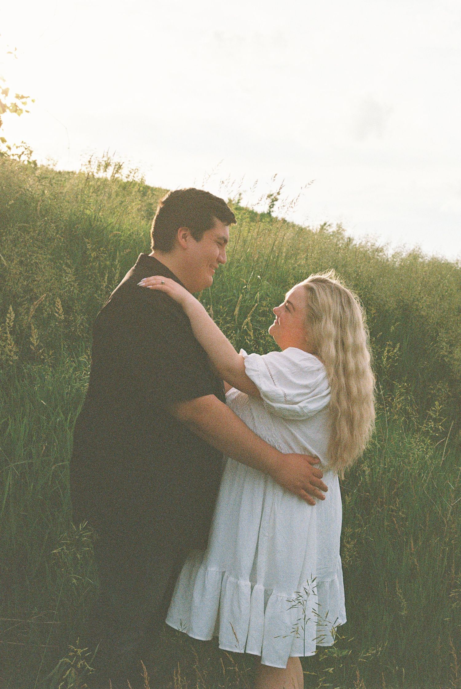 A couple embraces in a lush green field at golden hour, the woman in white dress smiling up at the man in black.