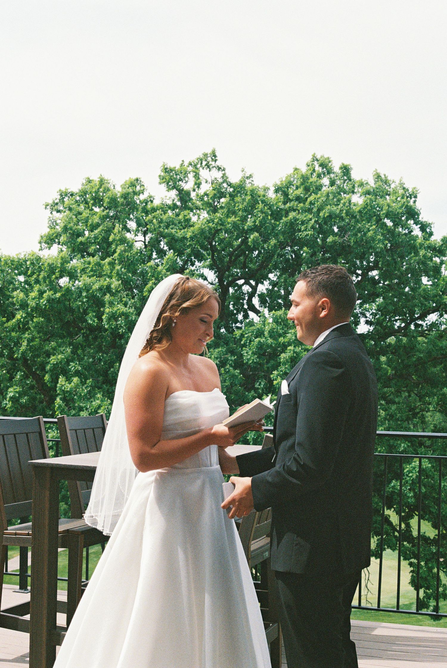 Bride and groom exchange vows on outdoor deck surrounded by lush green trees during wedding ceremony.