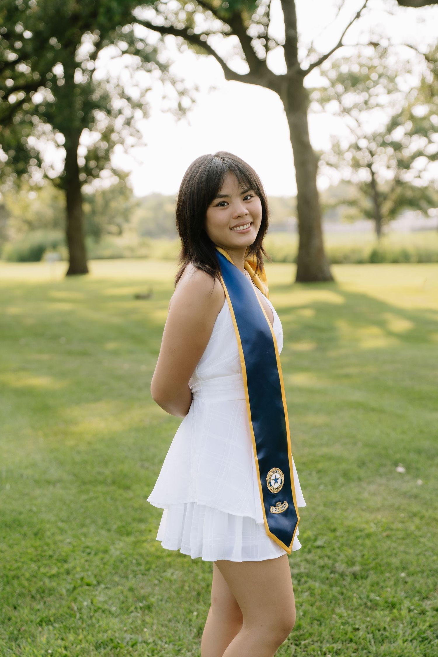 Smiling young woman in white dress and navy graduation sash poses outdoors in green park setting.