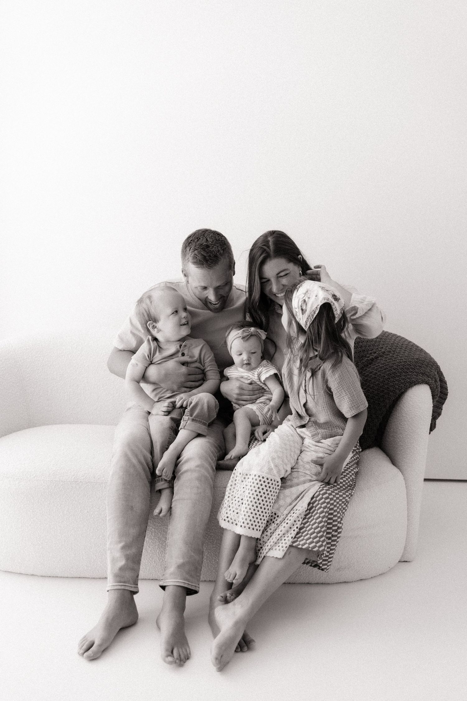 Black and white family portrait of parents with two toddlers and a baby sitting together on a white sofa.