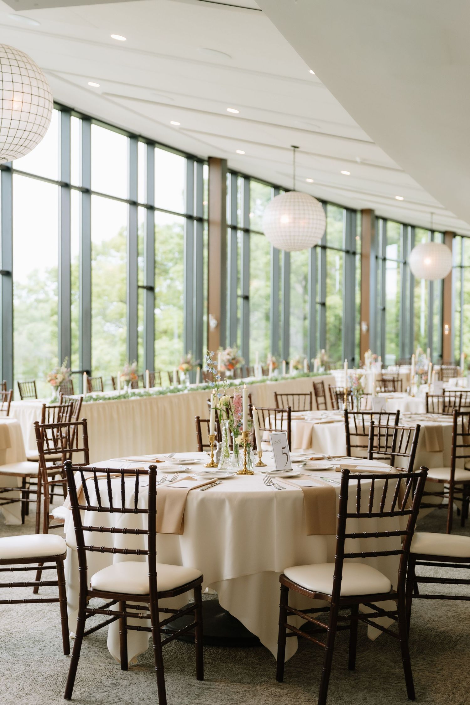 Elegant wedding reception hall with round tables, chiavari chairs, cream linens, and floor-to-ceiling windows overlooking greenery.