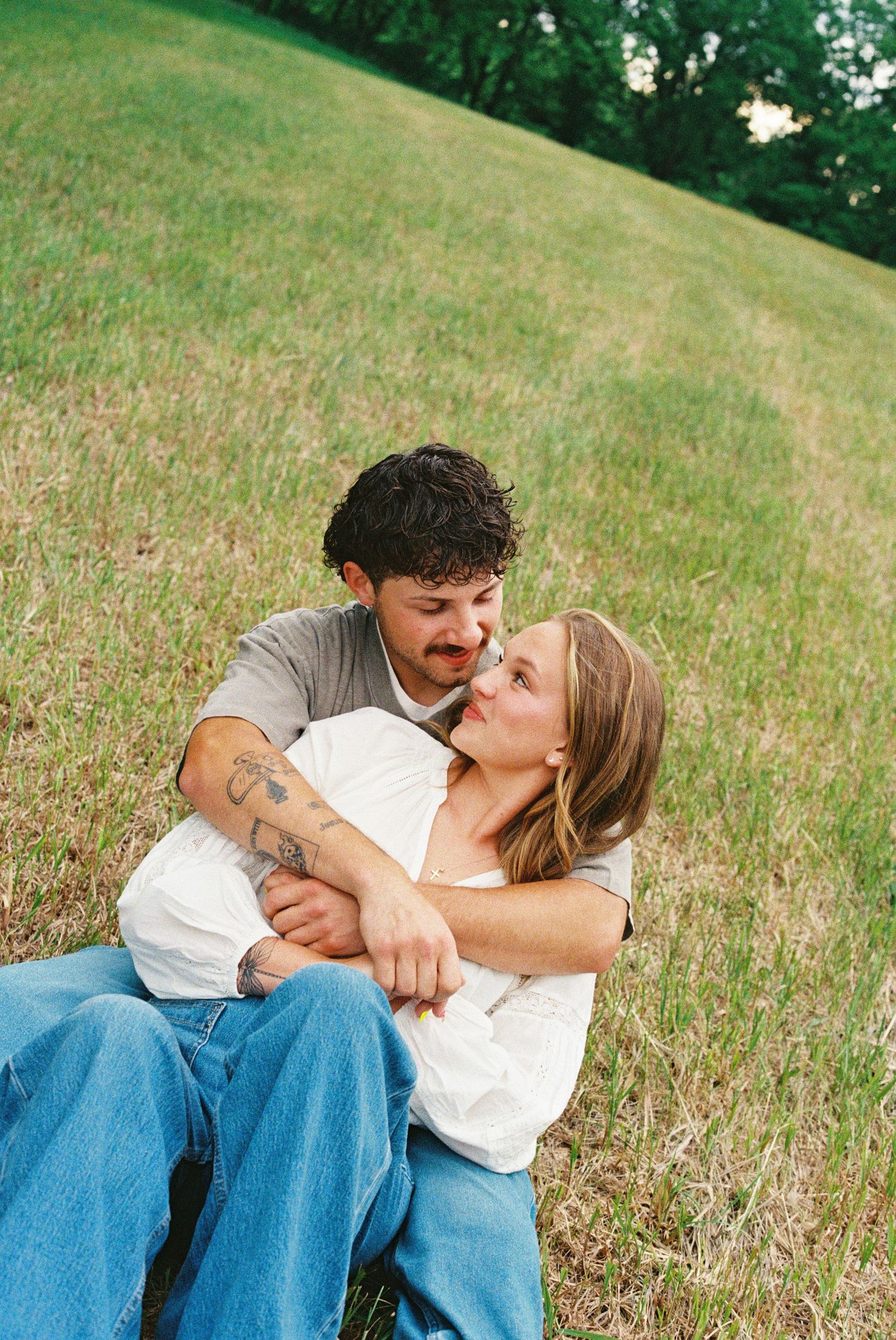 A couple sits together on a grassy hill, embracing and sharing a tender moment outdoors.