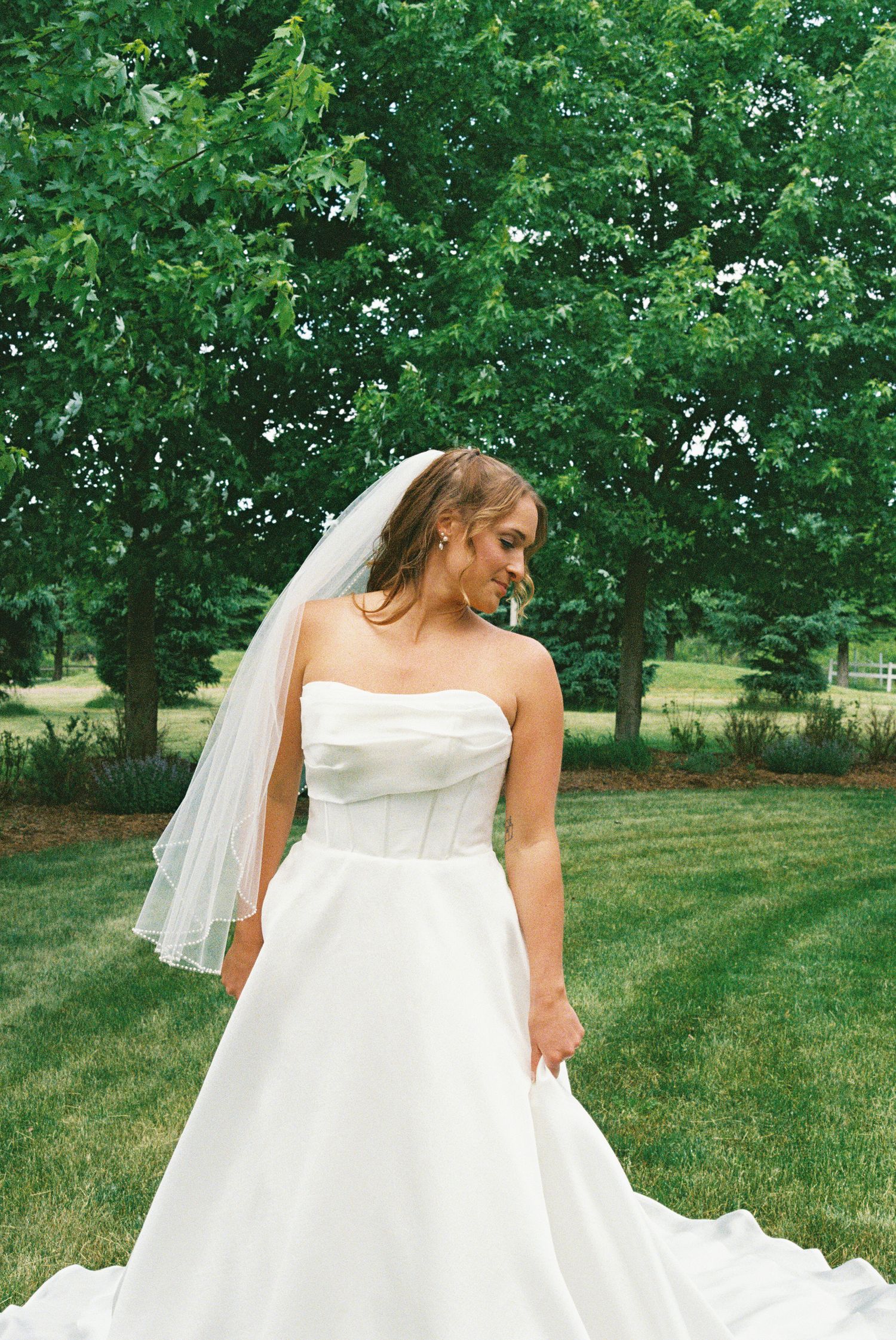 Bride in strapless white ball gown with veil poses outdoors on green lawn with lush trees in background.