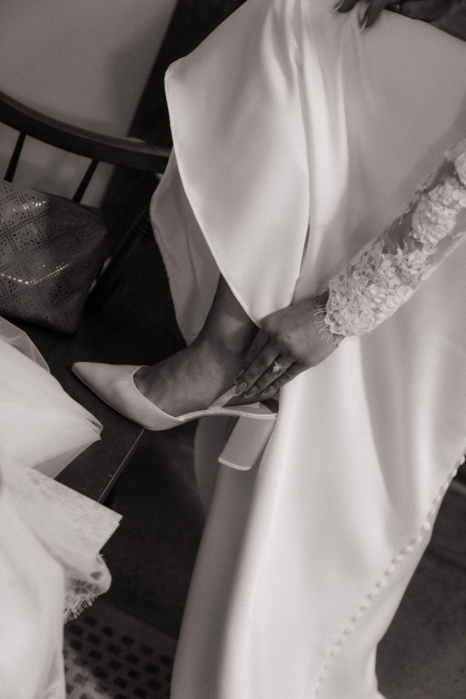 Black and white photo of a bride in a white lace wedding dress putting on elegant heeled bridal shoes.