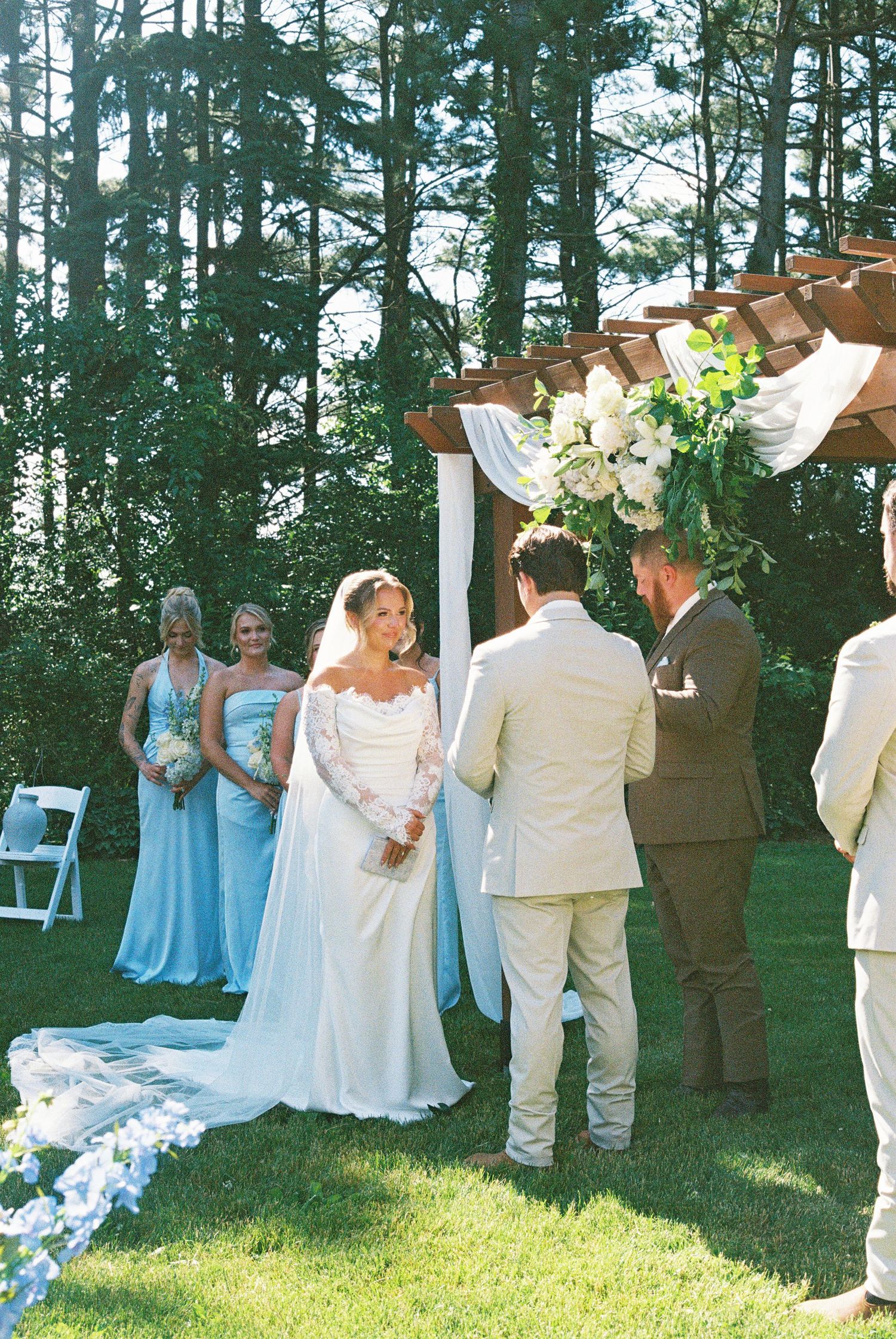 Bride in white gown with veil exchanges vows with groom outdoors, bridesmaids in blue dresses beside floral arch.