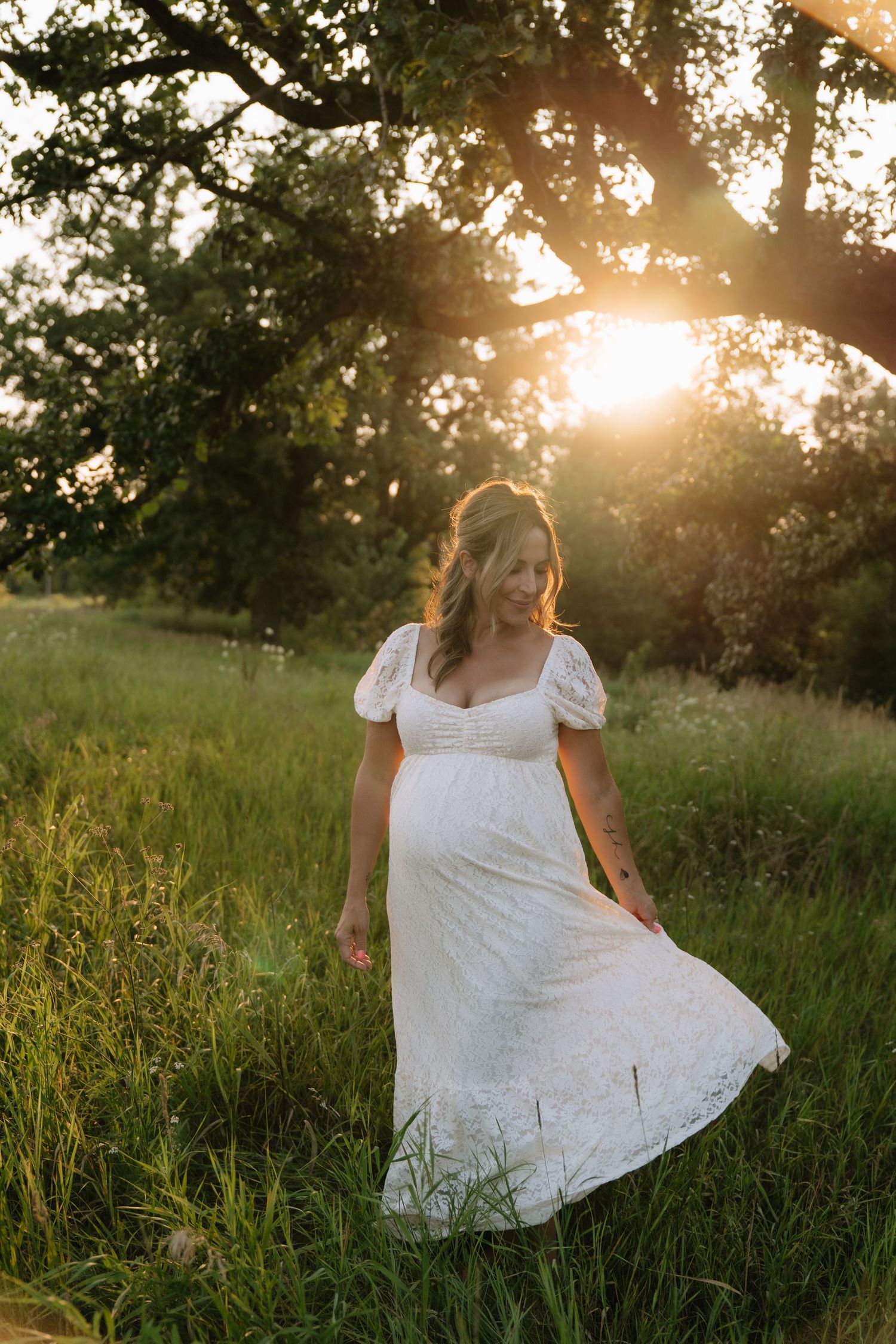 Pregnant woman in white lace dress walking through green meadow at golden sunset.