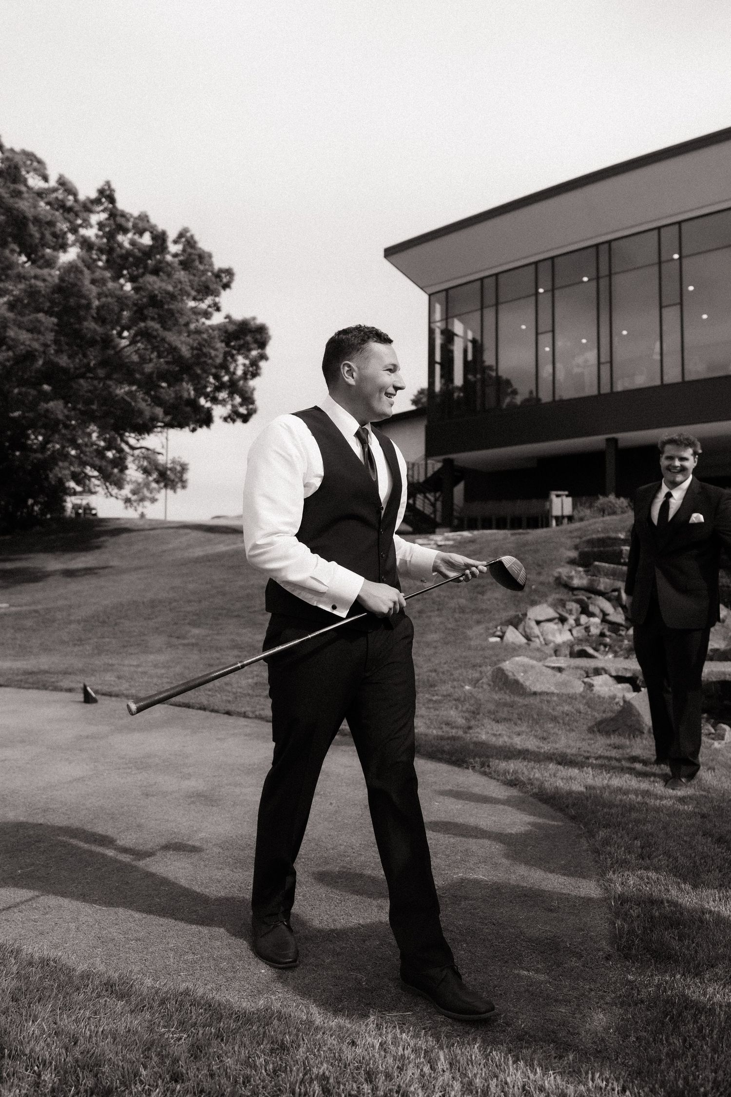 Black and white photo of a smiling groom holding a golf club outdoors near a modern building.