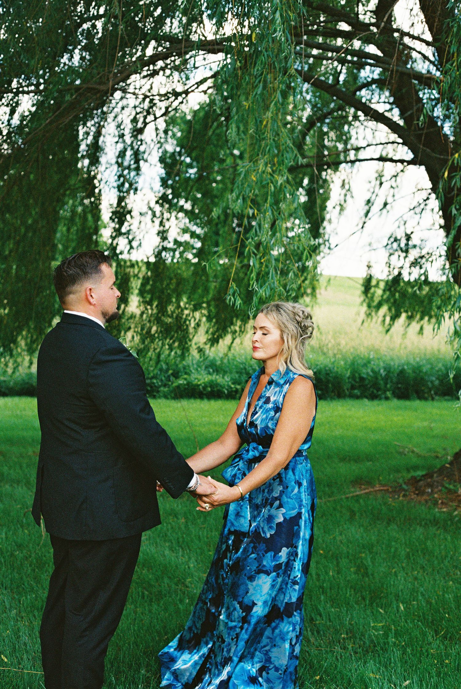 Couple holding hands under a weeping willow tree, man in black suit and woman in blue floral gown, outdoor engagement photo.