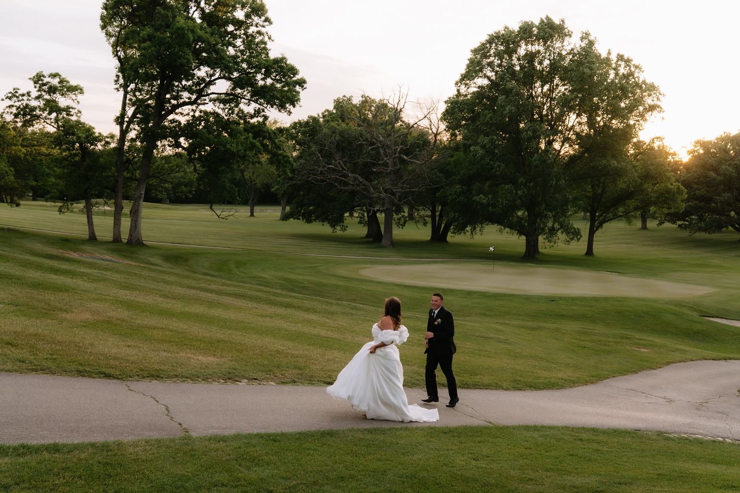 Bride and groom walking on a golf course at sunset, surrounded by lush green trees and grass.