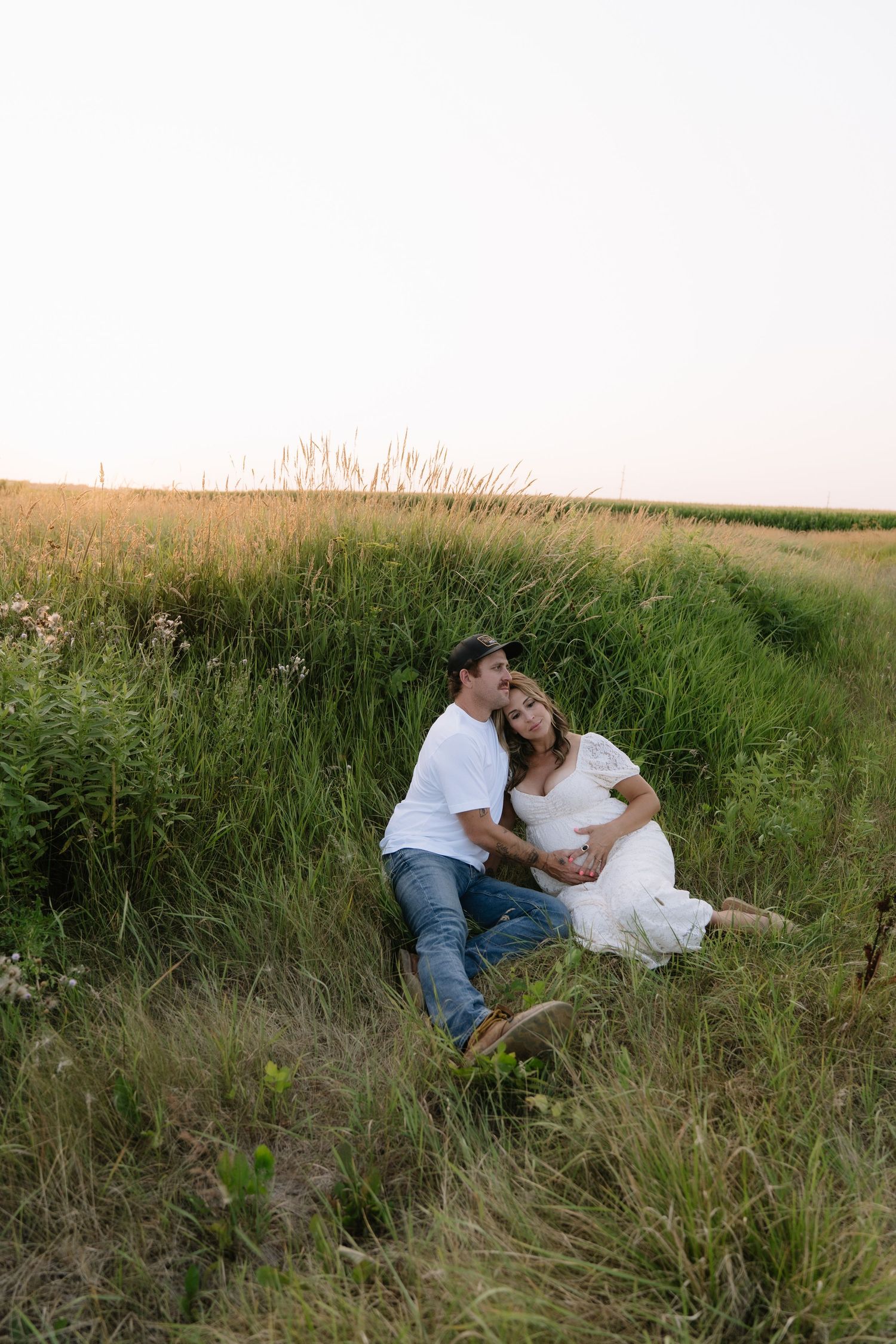 A couple sits together in a lush green field at sunset, the woman in white leaning on her partner.