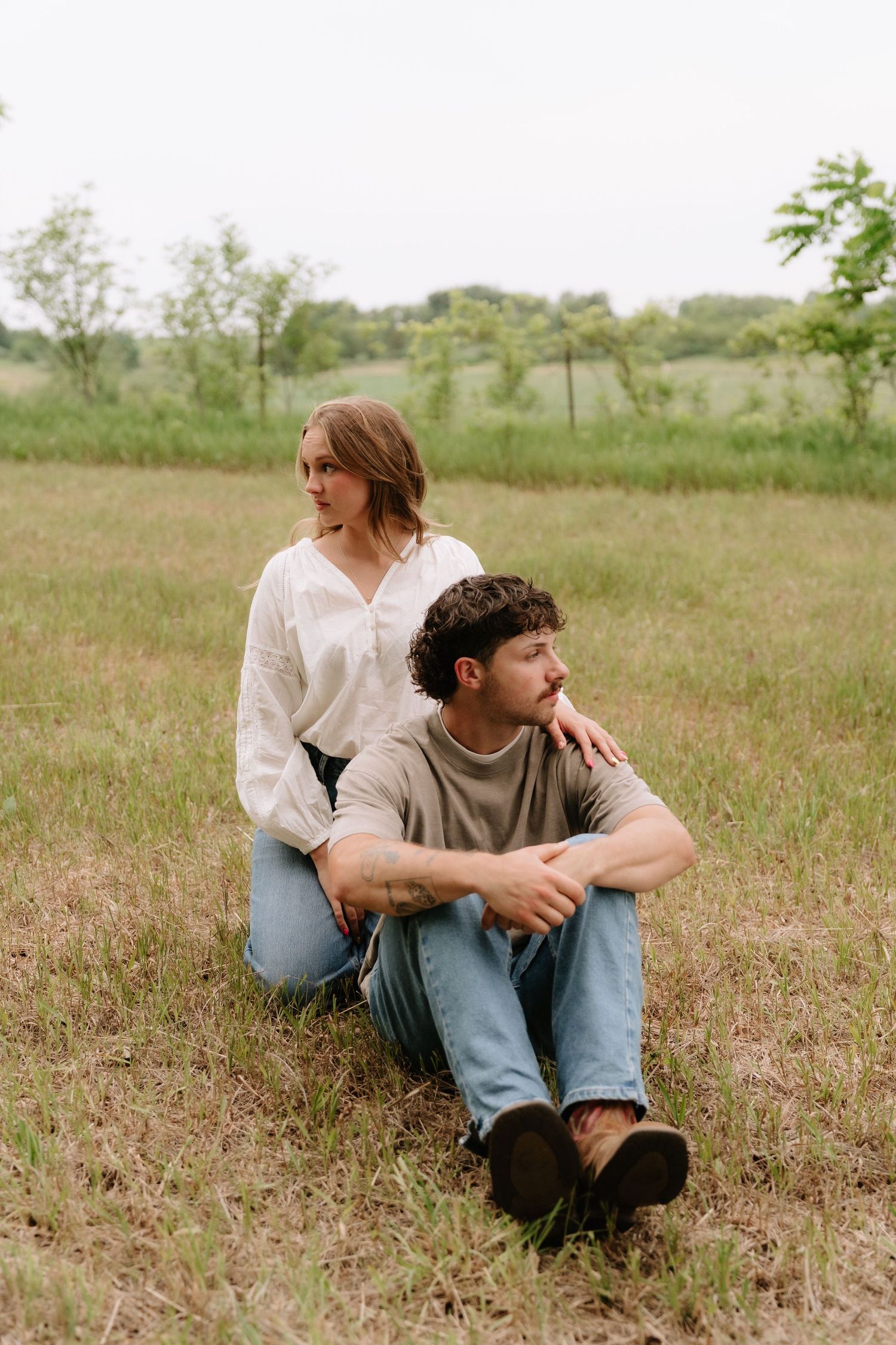 Couple sitting together in a grassy field, woman in white blouse resting hand on man's shoulder.