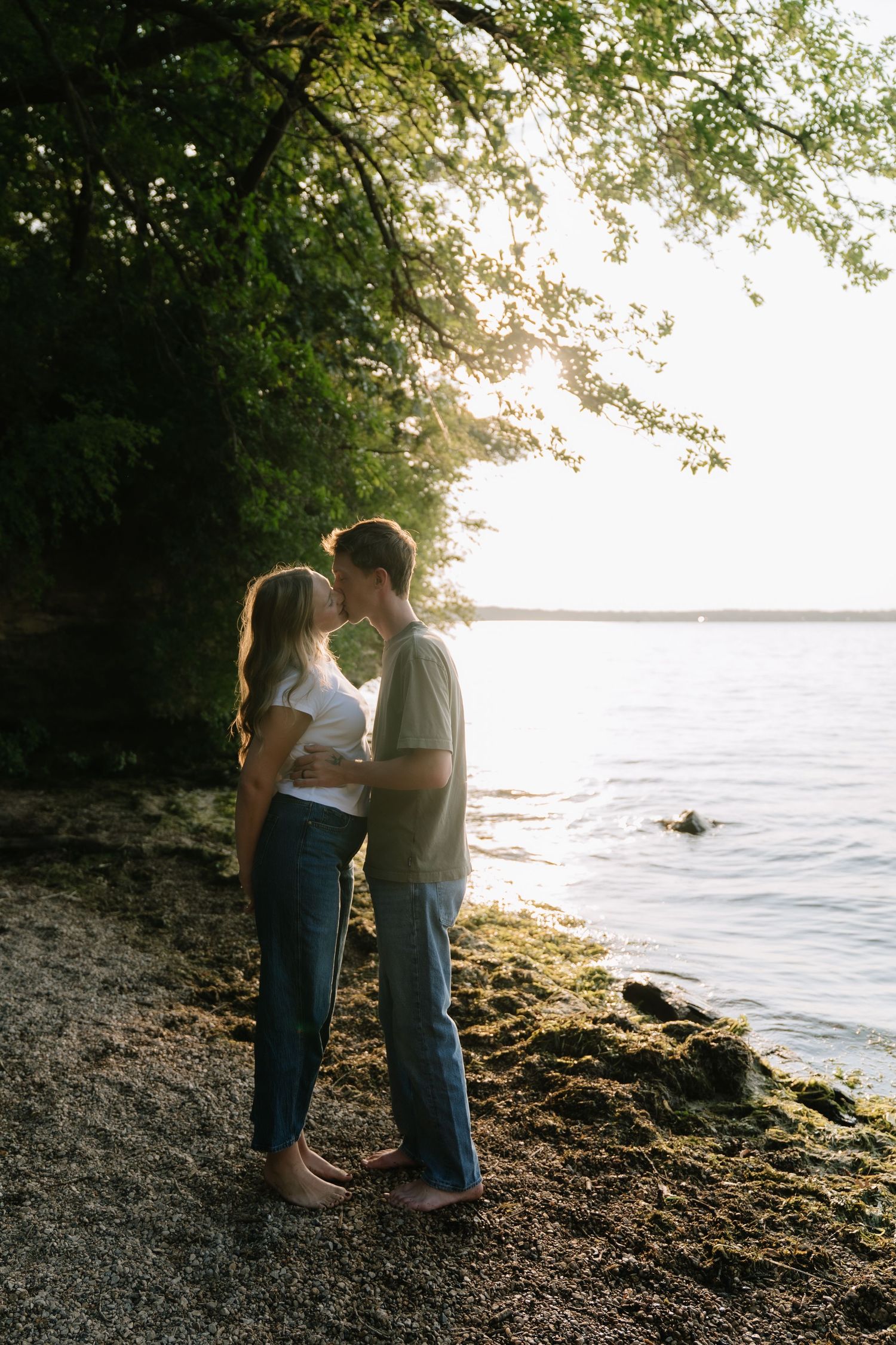 A couple shares a romantic kiss by a sunlit lakeside shore, surrounded by lush green trees at golden hour.