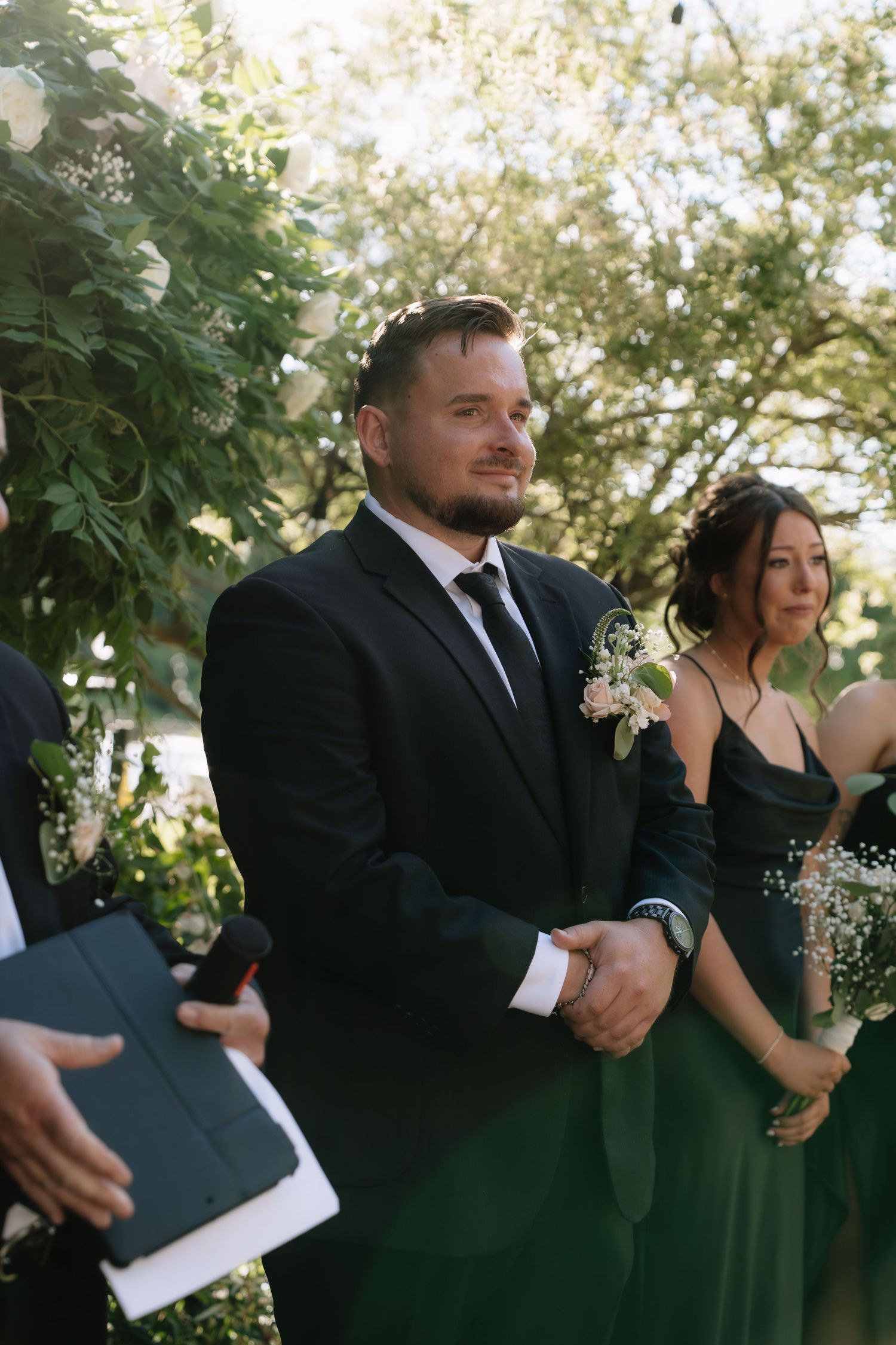 Groom in black suit with boutonniere smiling at outdoor wedding ceremony, bridesmaid in green dress beside him.